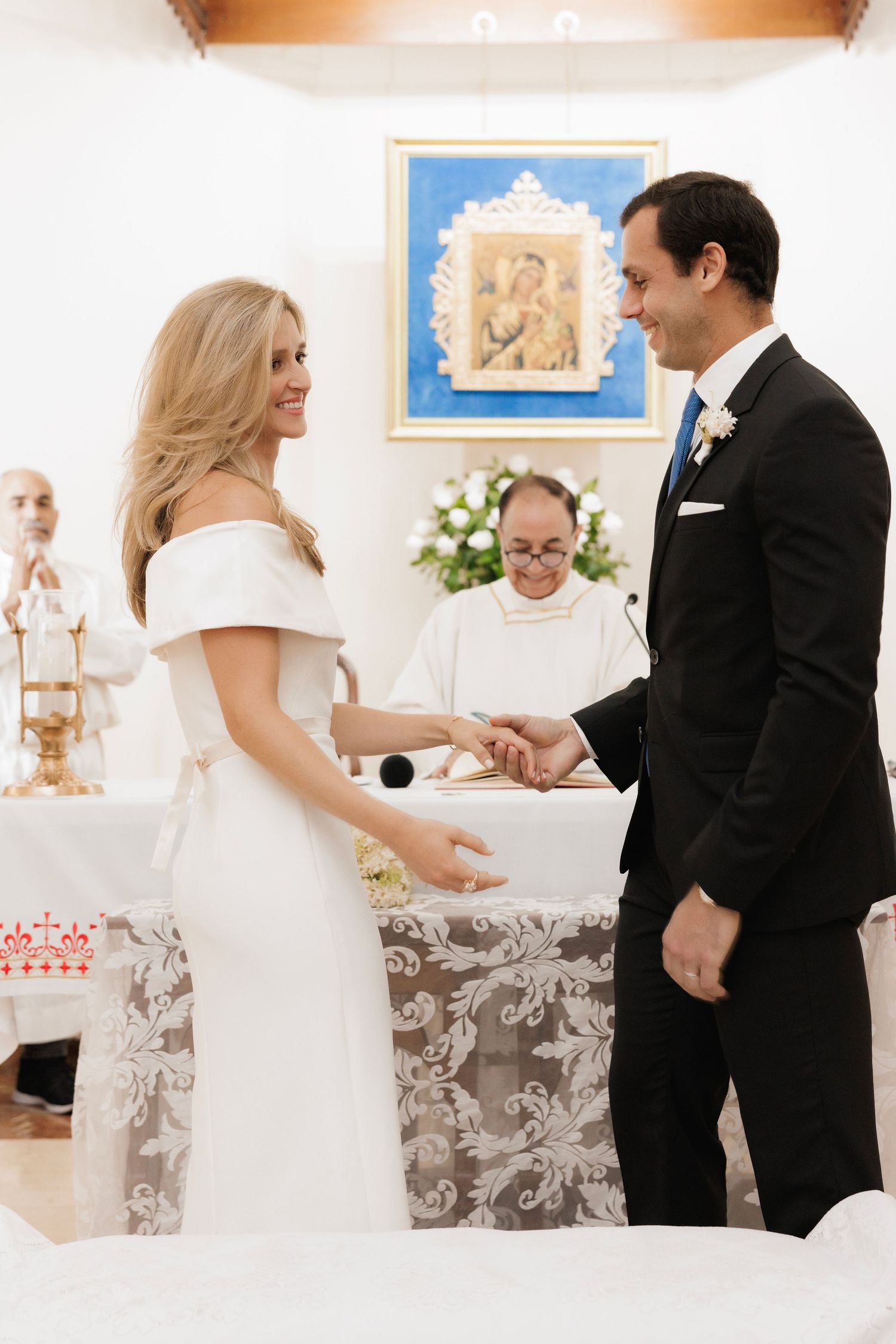 A couple in wedding attire exchanges vows during an intimate indoor ceremony with floral decorations and artwork.