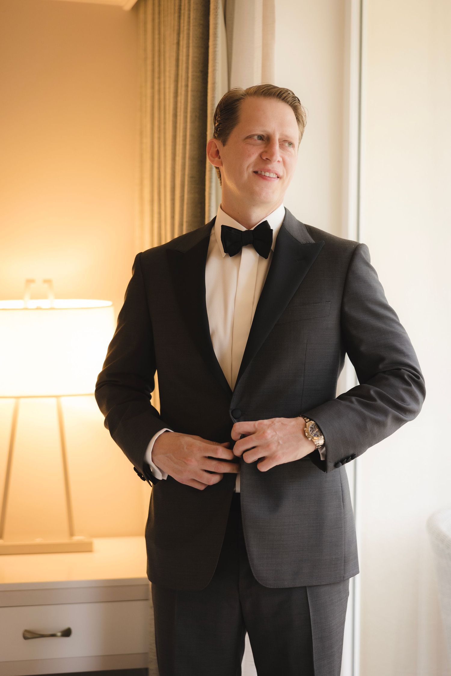 A groom in a classic black tuxedo and bow tie adjusts his jacket while standing by a window with warm lighting.