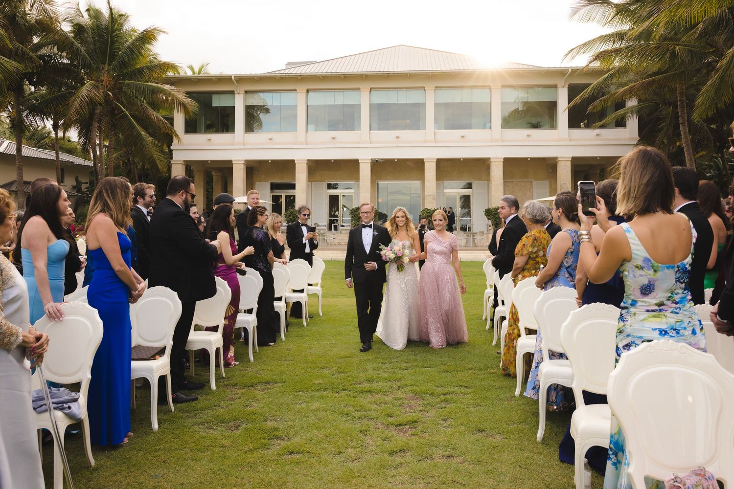 An outdoor wedding ceremony setup on a lawn with white chairs and palm trees in front of an elegant plantation-style venue.