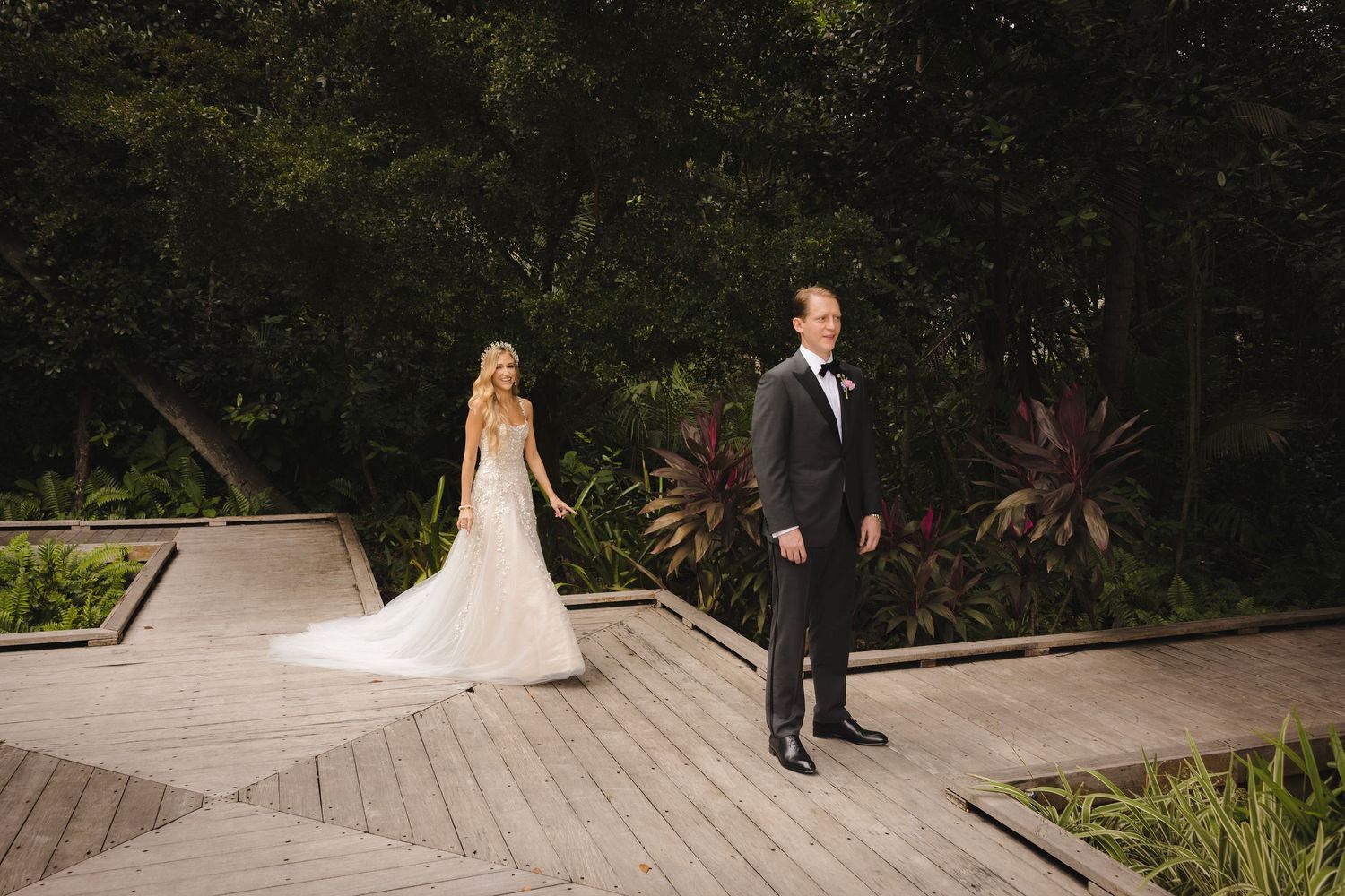 A couple in wedding attire walks along a modern stone pathway surrounded by lush tropical landscaping.
