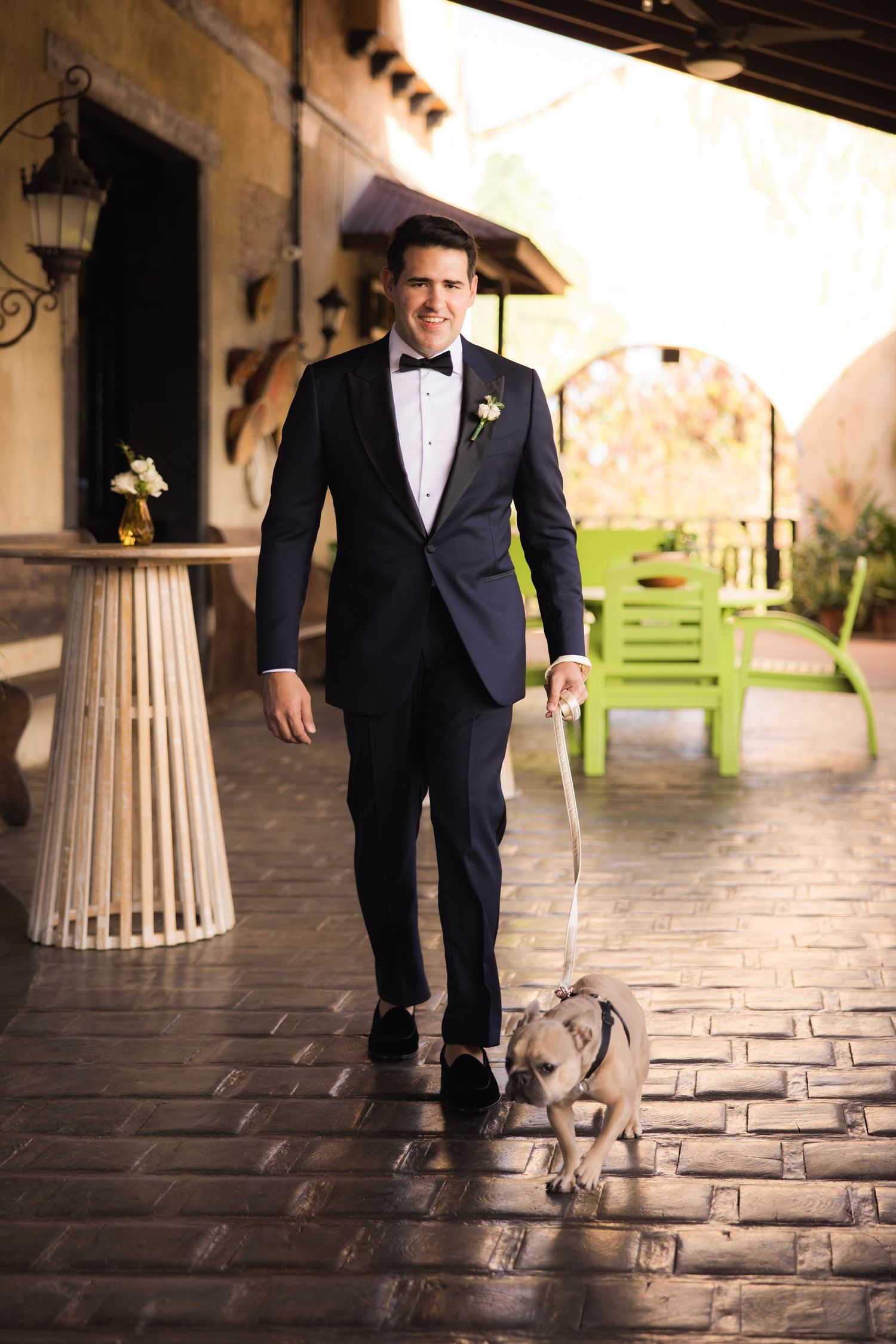 A person in a black tuxedo walks with a small dog on a tiled patio area.