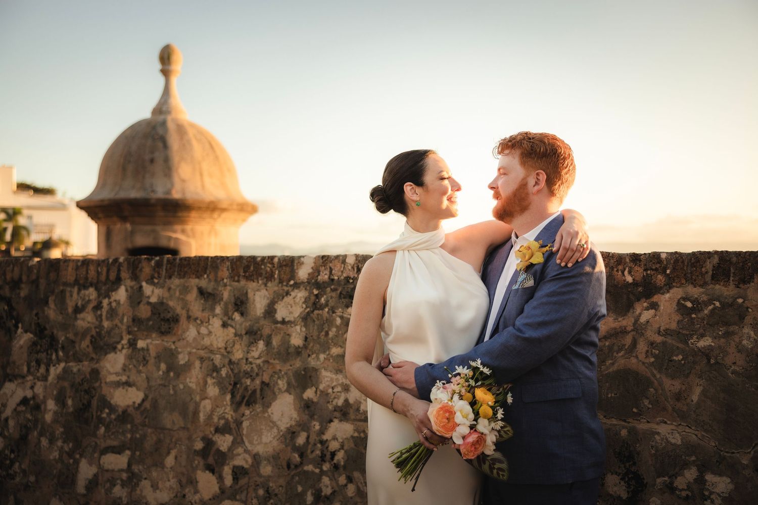 Romantic couple embraces against stone wall at sunset creating intimate silhouette with floral bouquet.