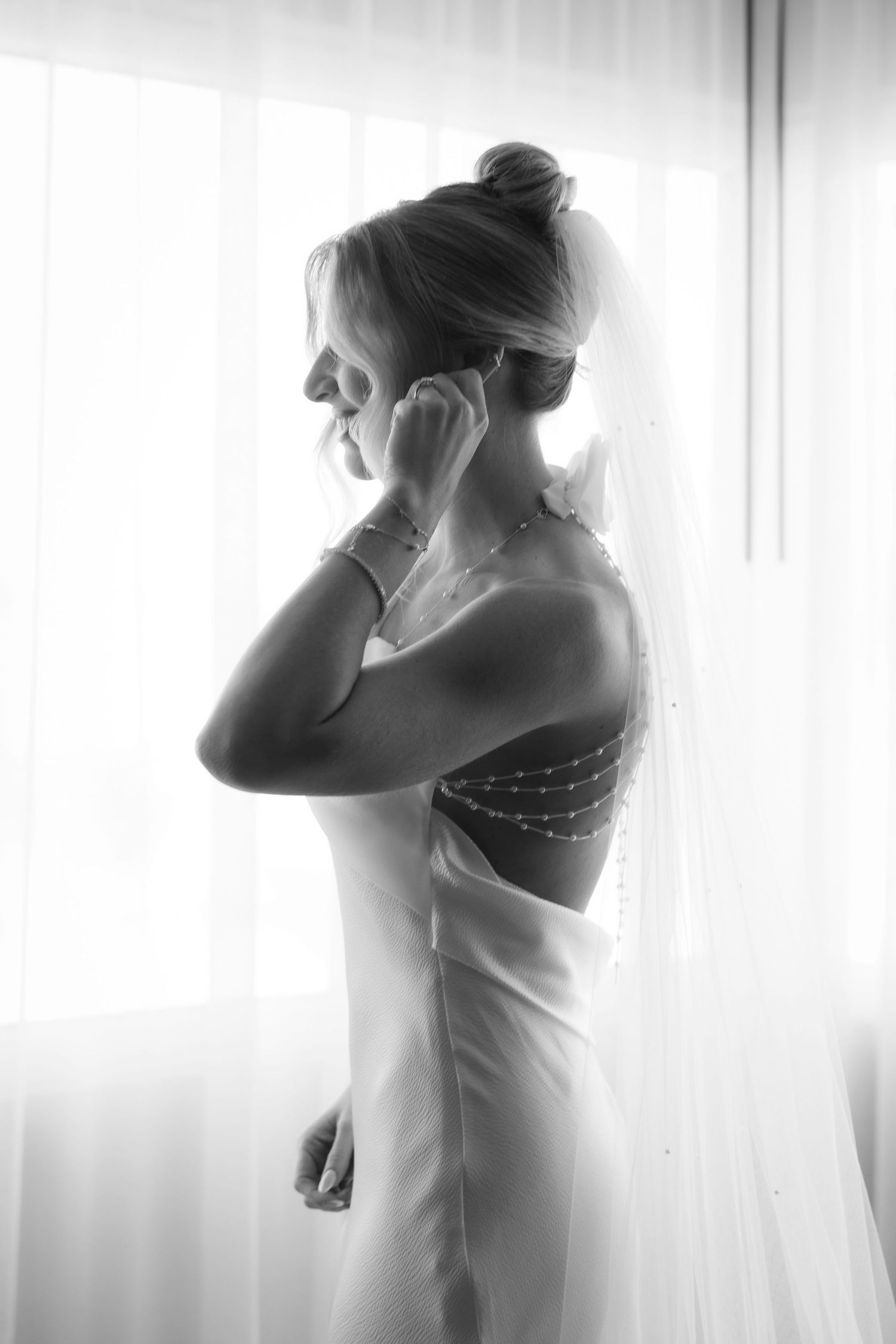 A black and white silhouette of a bride in her wedding dress standing by a window in soft natural light.
