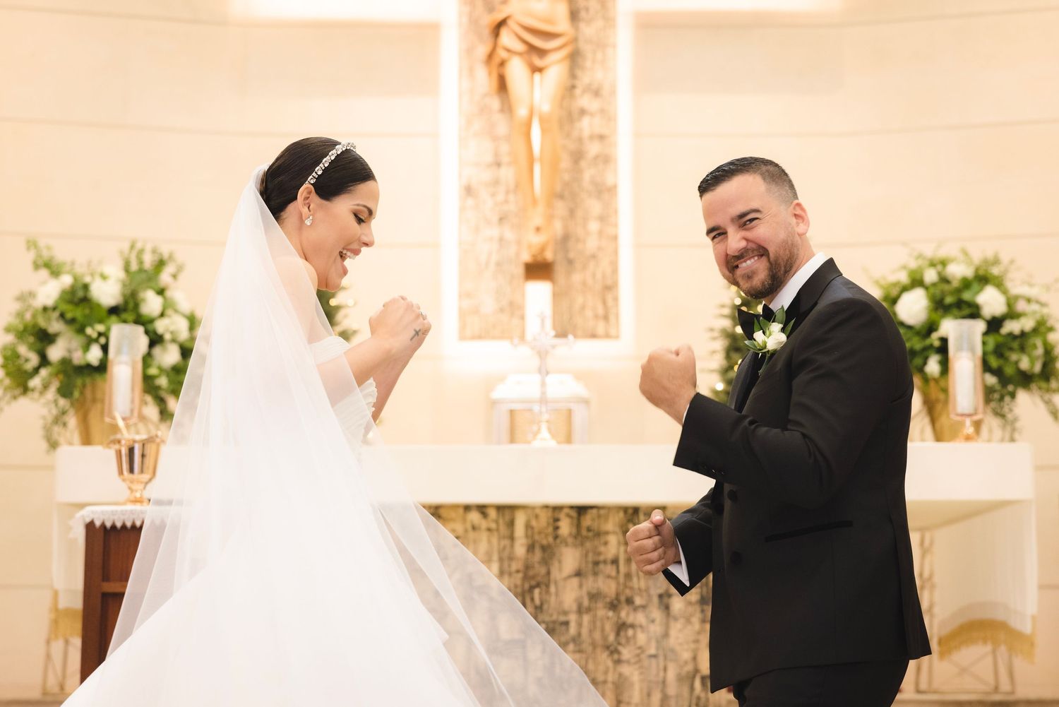 A joyful moment at a wedding ceremony with bride and groom sharing laughter in front of a church altar with floral decor.