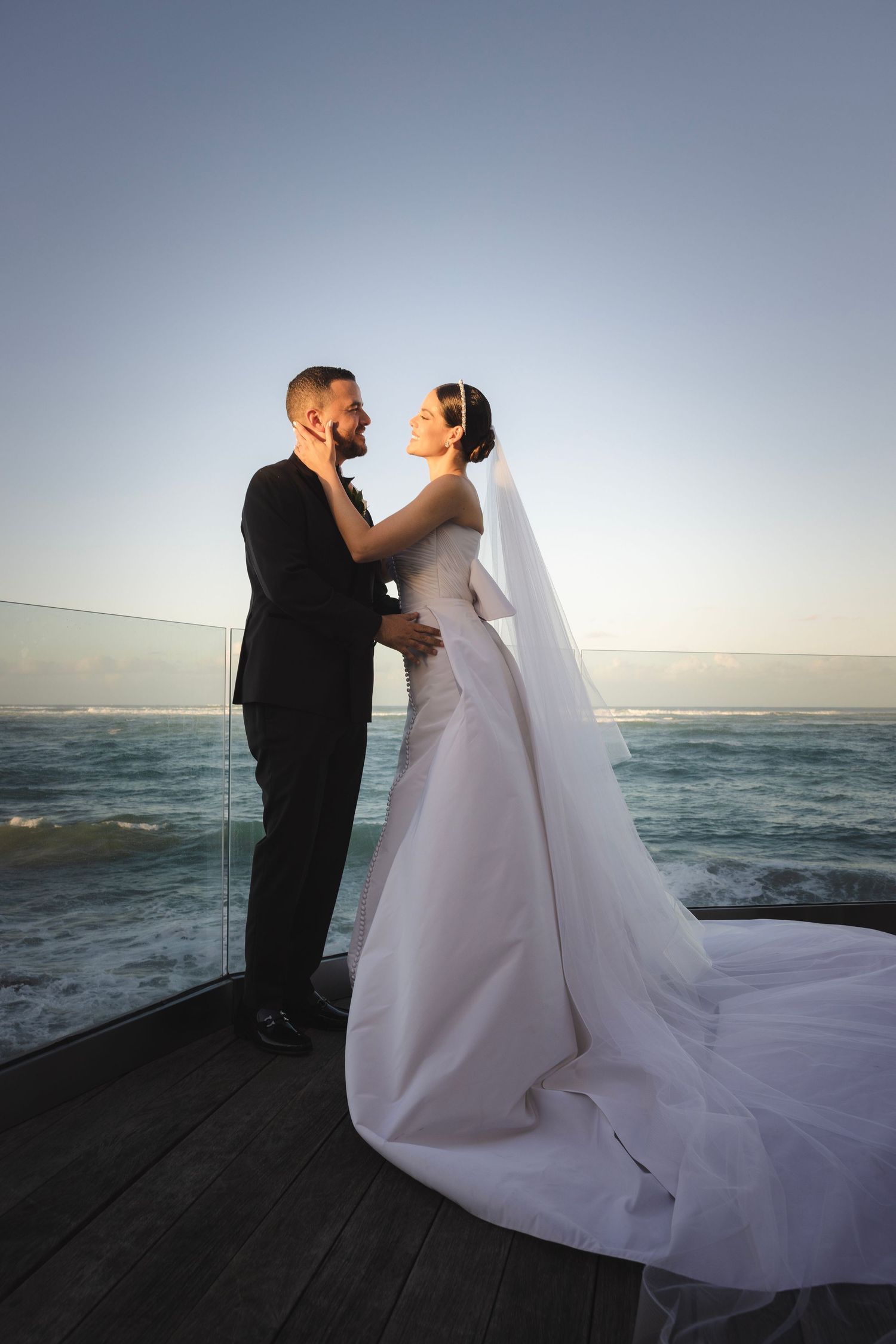 A bride and groom share a romantic moment on a wooden deck overlooking the ocean at sunset.