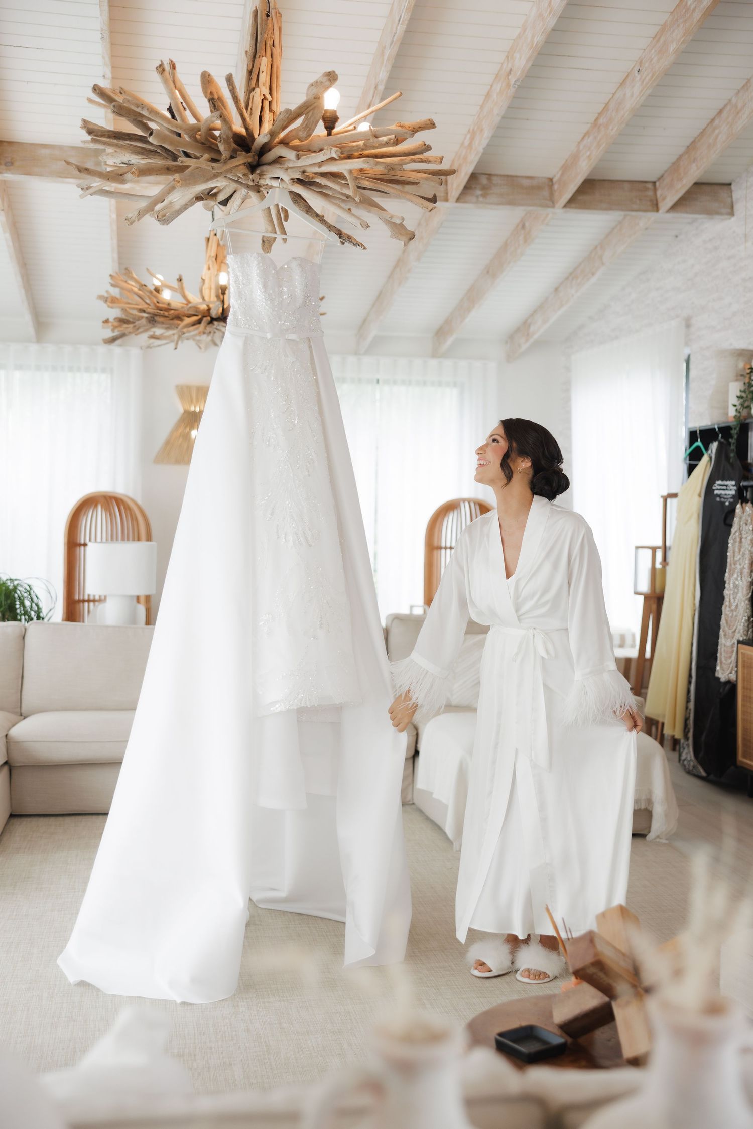 A white wedding dress hangs from a driftwood chandelier in a bright modern room with arched windows.