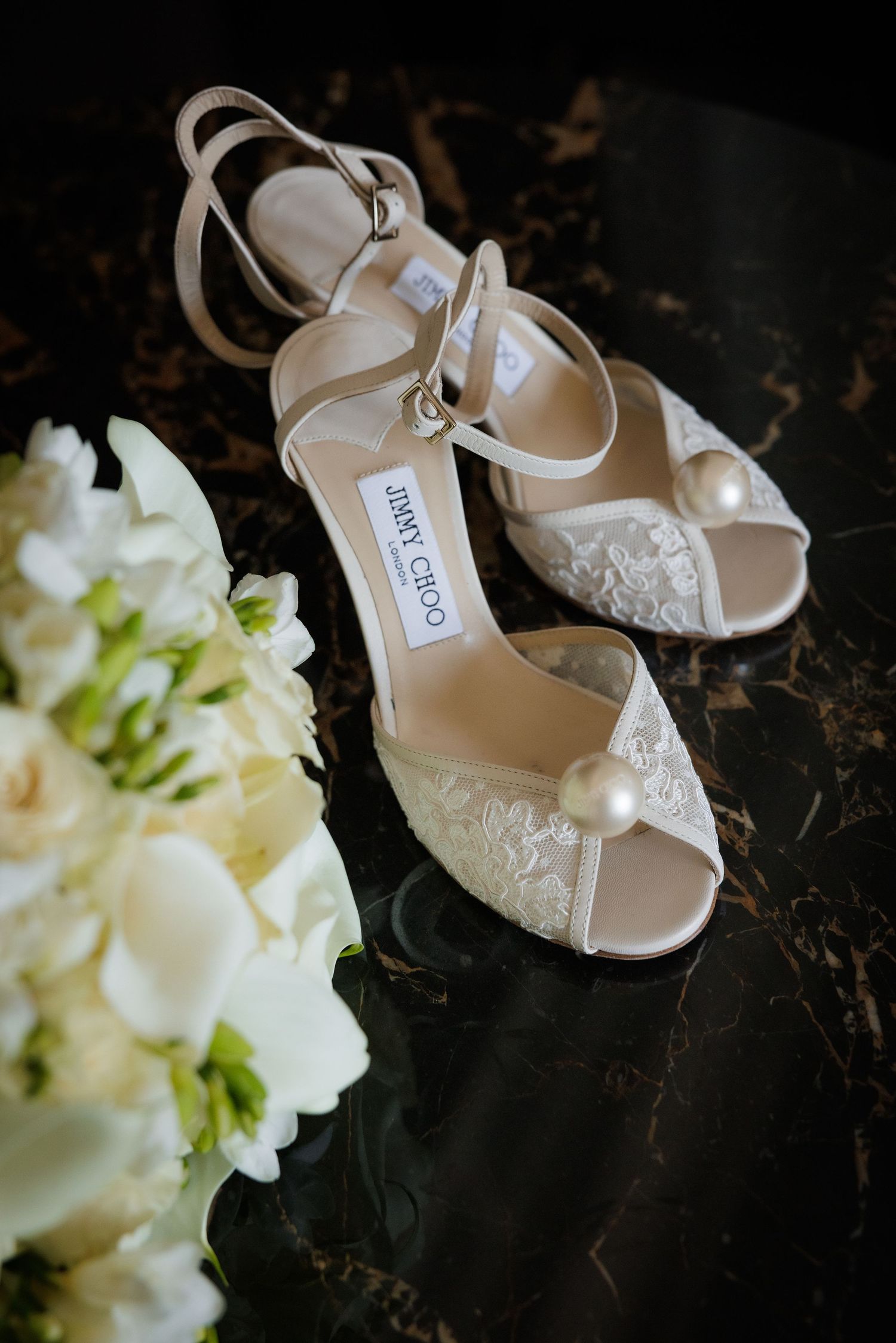 Elegant white lace bridal shoes paired with a delicate white floral bouquet on a dark background.