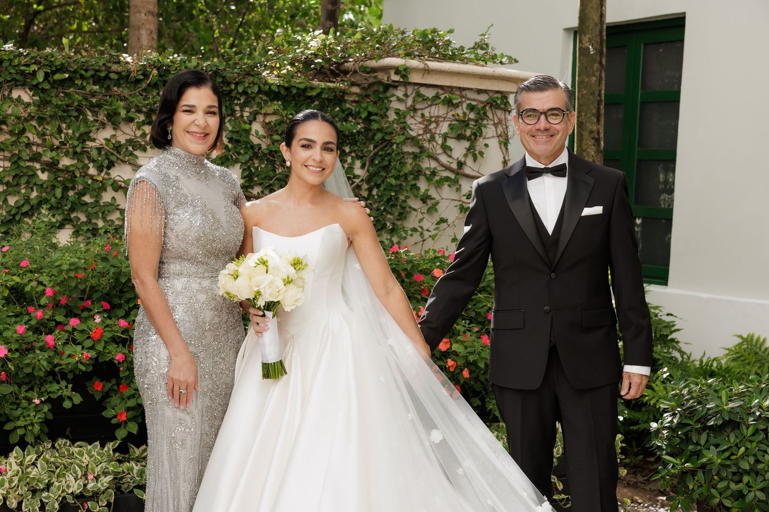 Wedding party poses in front of white building with lush green garden backdrop.