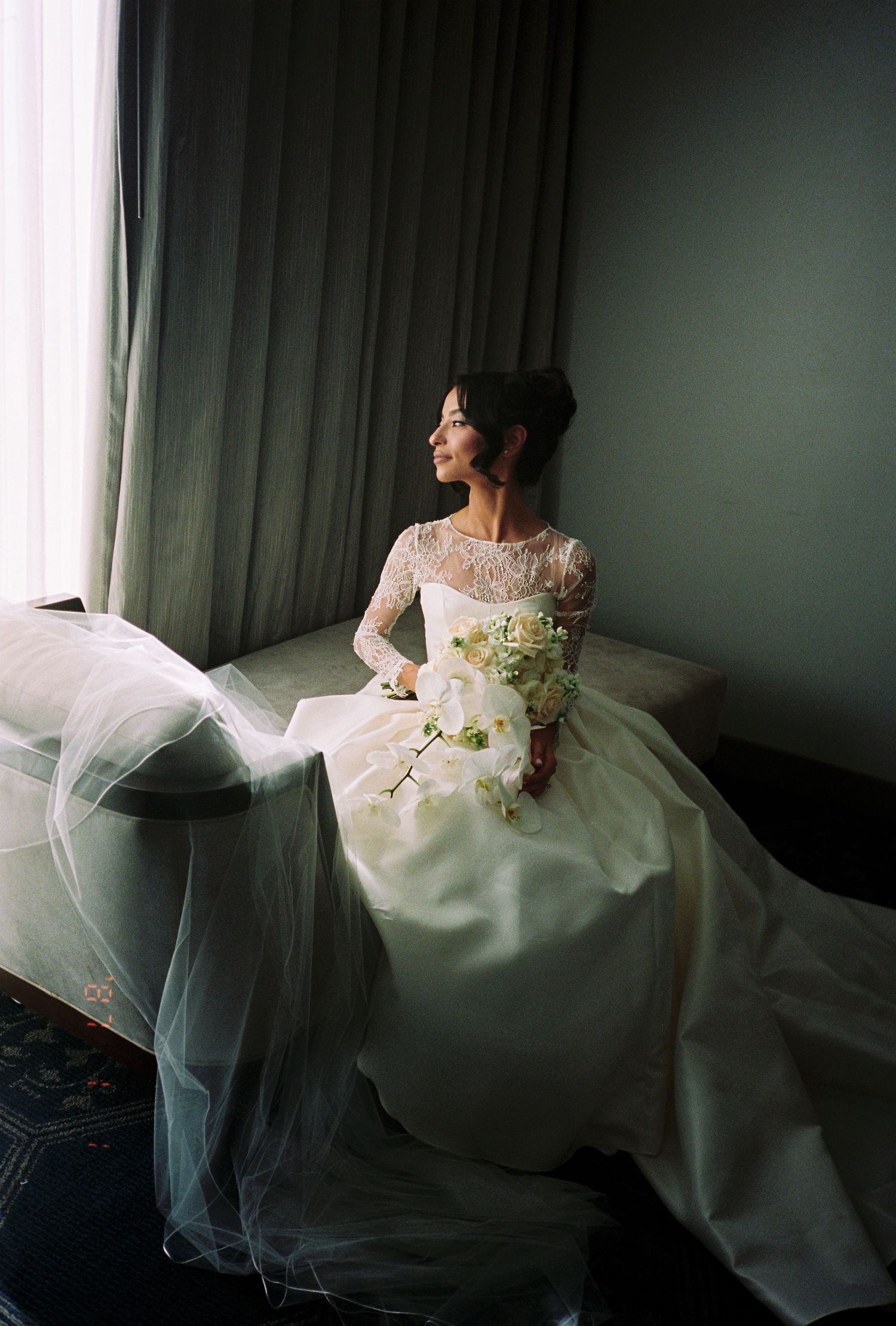 Bride in lace wedding gown with cathedral veil sitting by window holding white floral bouquet.