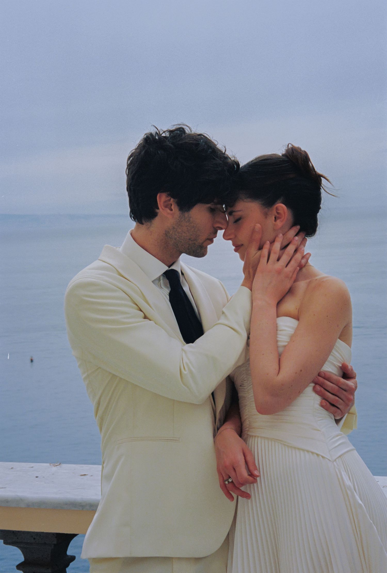 A couple in wedding attire share a romantic moment by the sea, the groom in cream suit, bride in white dress.