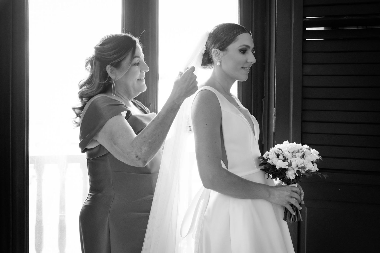 Black and white photo showing final wedding dress preparations by a window with soft natural light.