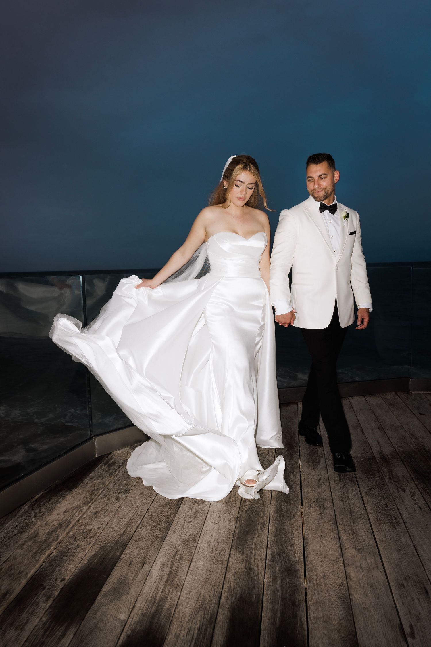 A couple in wedding attire poses playfully on a rustic wooden deck at dusk.