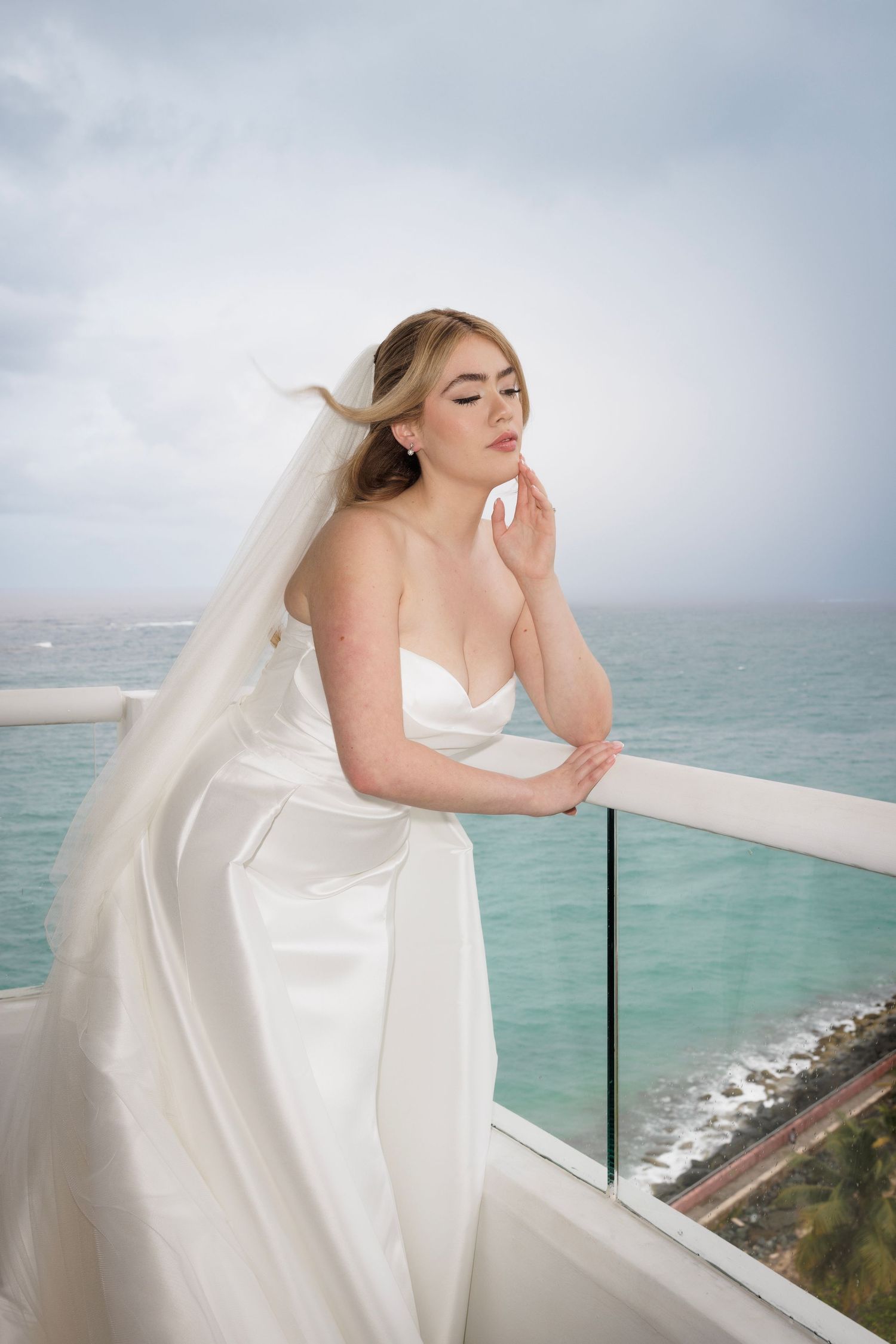 Bride in white dress standing on oceanfront balcony with flowing veil in the breeze.