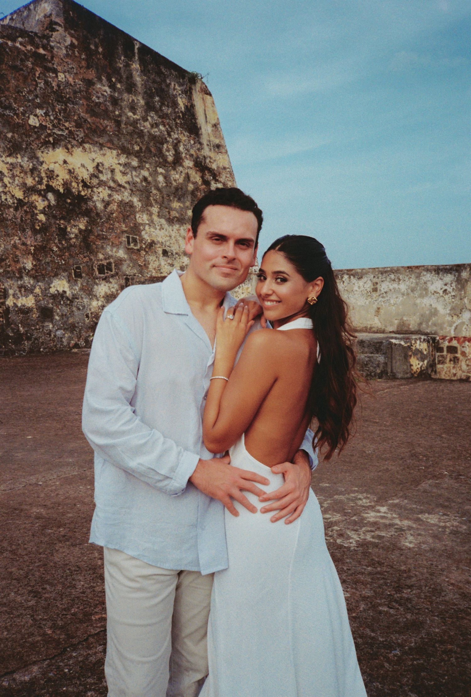 Couple in white outfits embracing at a historic stone fort during golden hour.