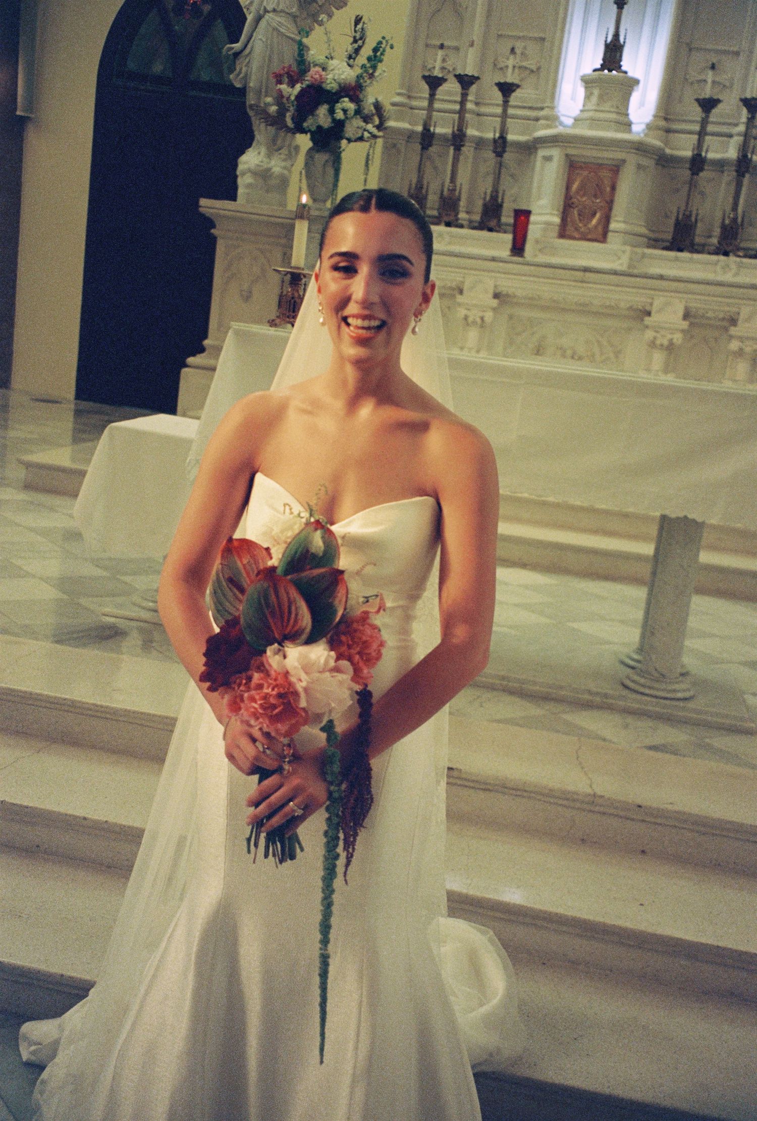 Smiling bride in strapless white gown holding tropical bouquet with pink peonies and red anthuriums at church altar.