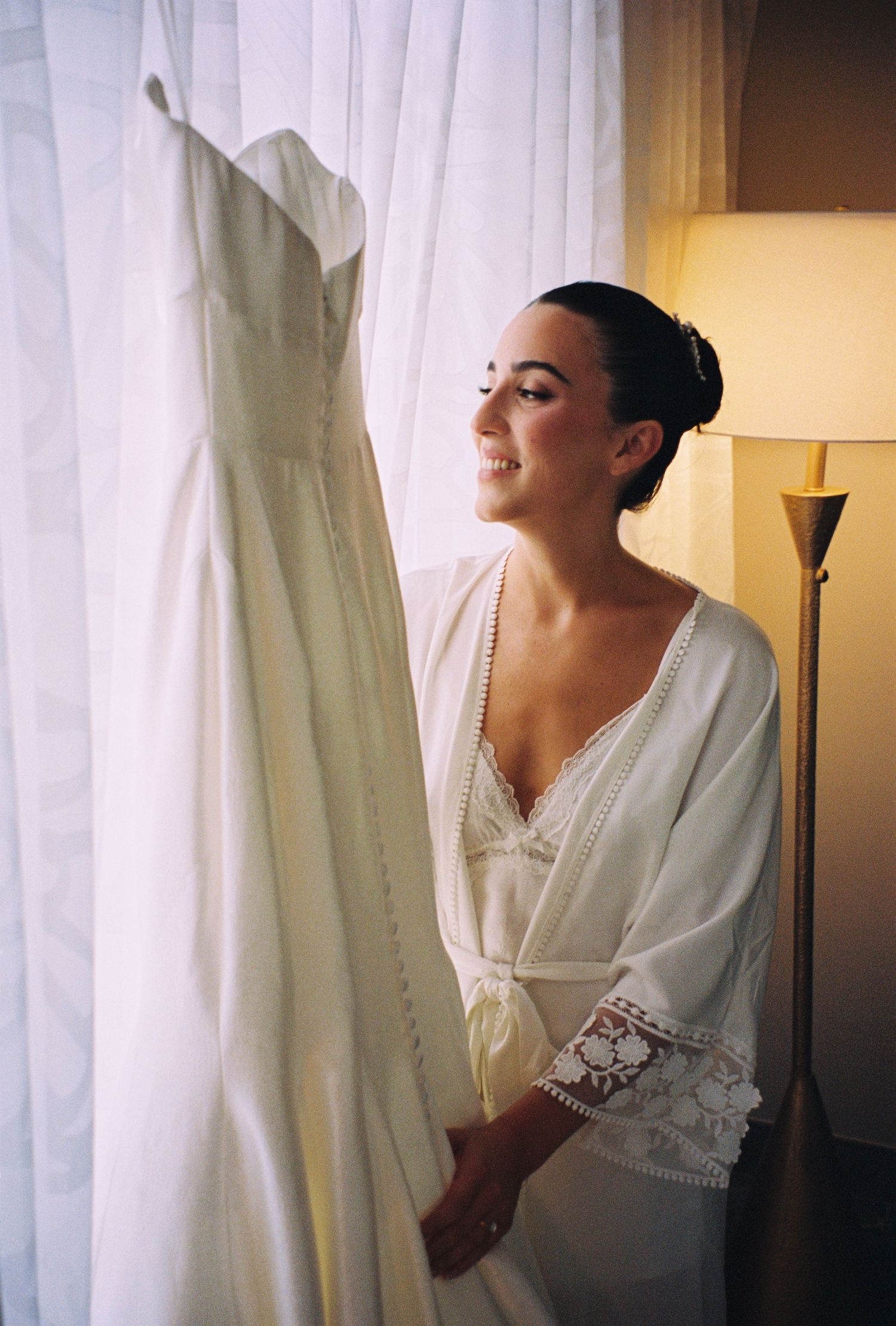 Bride in white lace robe admiring her wedding dress hanging by a curtained window with warm lamp light.