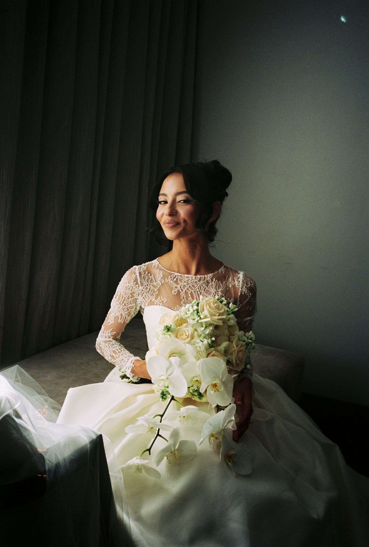 Bride in lace long-sleeve wedding gown holding white orchid bouquet, seated by window in soft natural light.