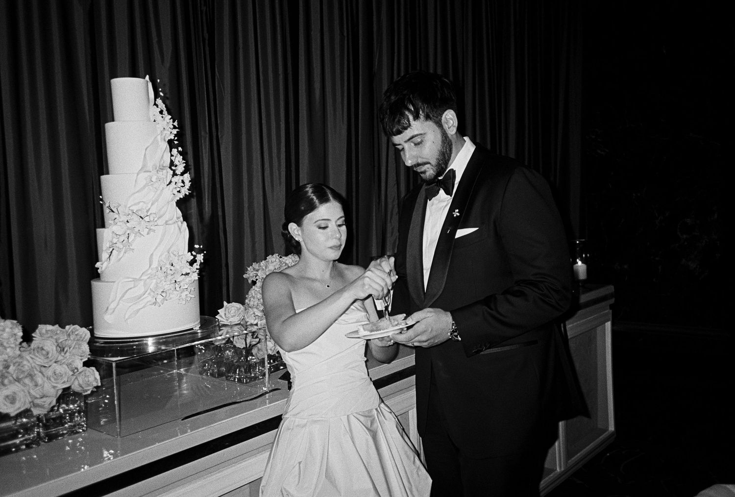Black and white photo of bride and groom cutting their elegant tiered wedding cake at a formal reception.
