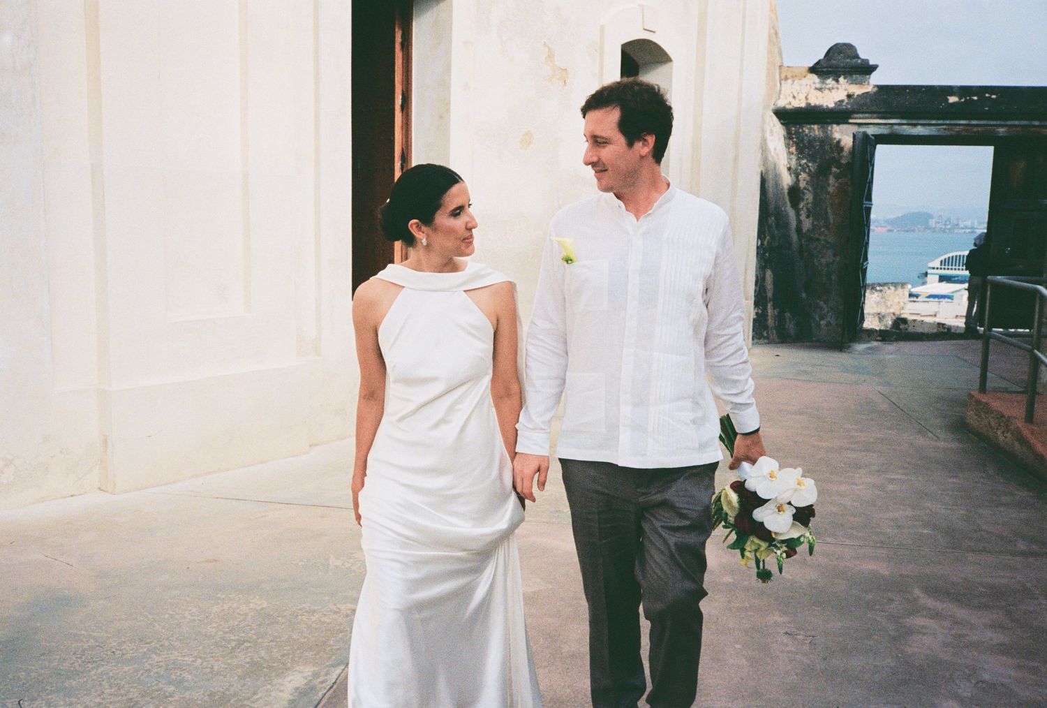 Bride and groom holding hands and smiling at each other on their wedding day in a tropical outdoor setting.