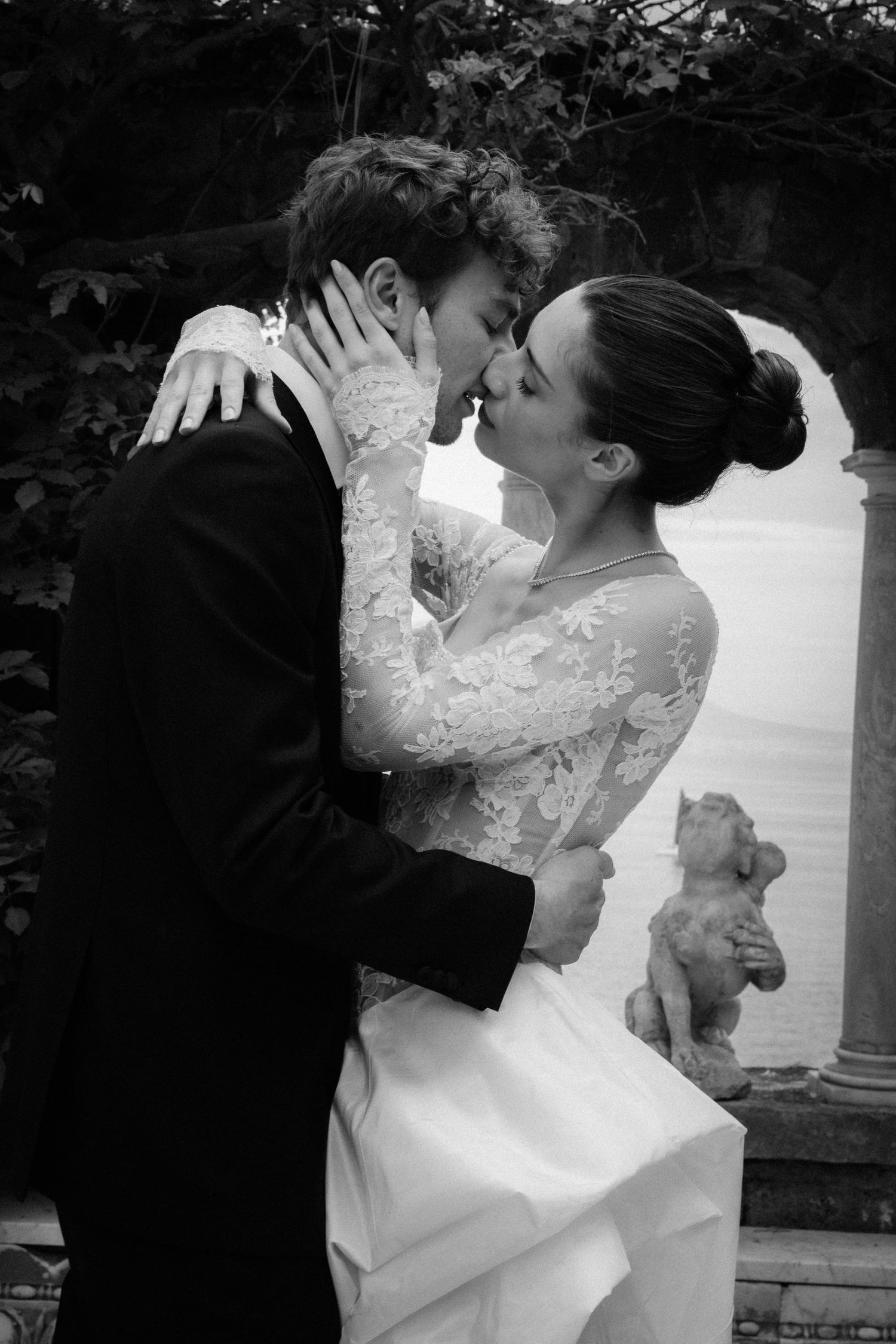 Black and white photo of a bride and groom sharing a romantic kiss in a garden, bride in lace gown.