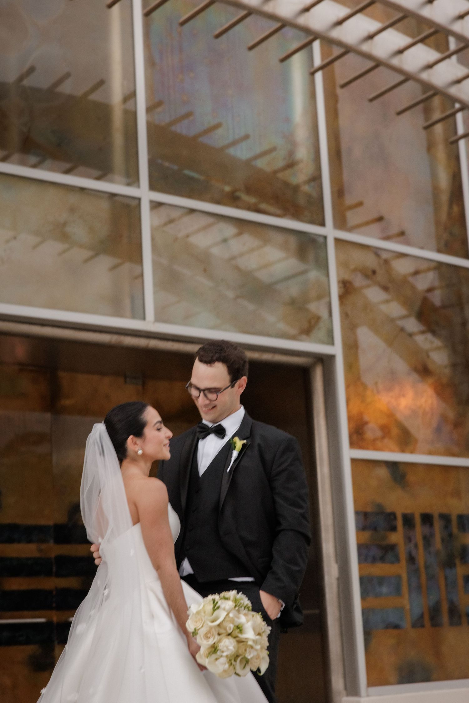 Wedding couple poses together in front of a modern building with glass windows and architectural details.
