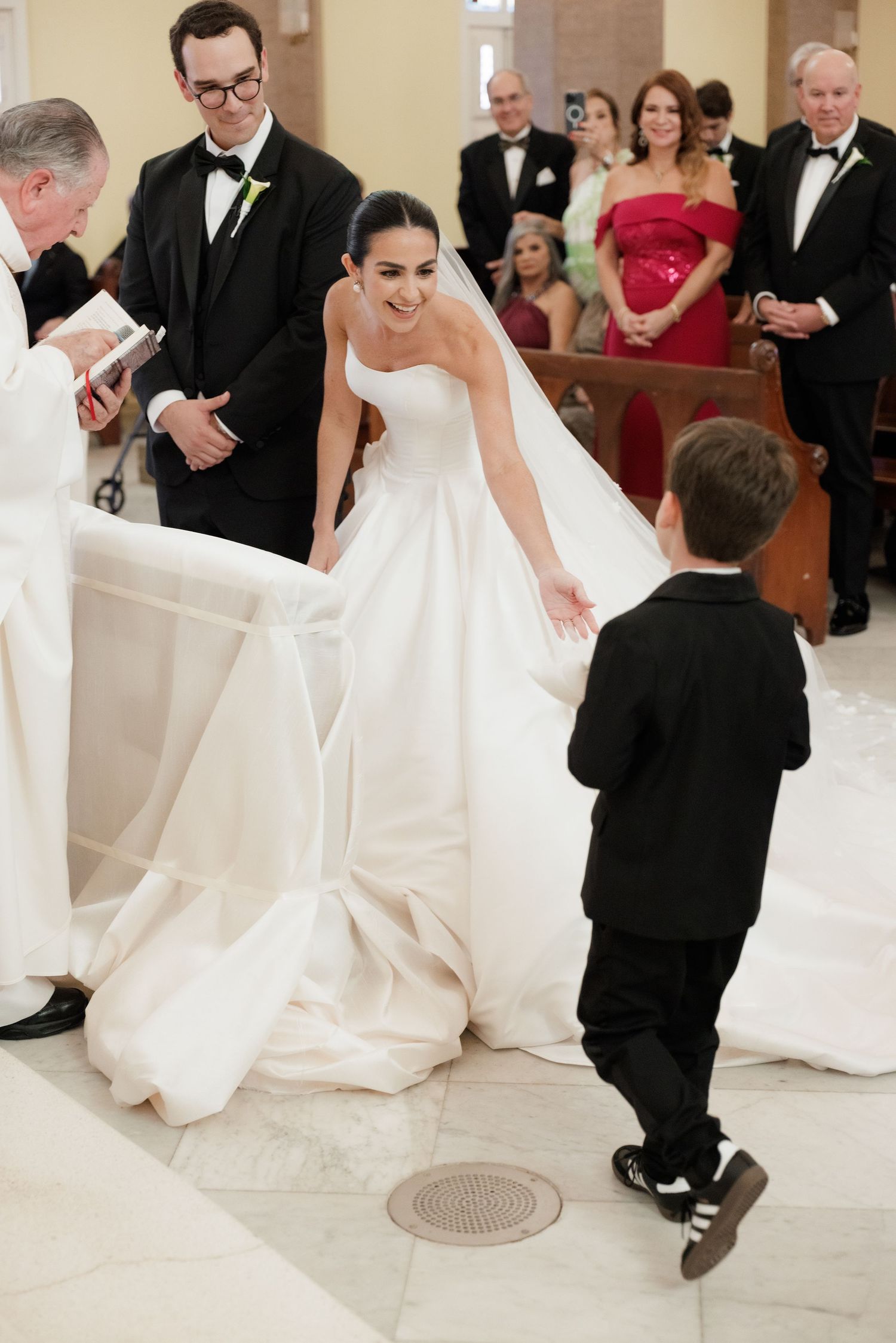 A wedding ceremony in progress shows attendees gathered in formal attire as a young boy approaches the bride's dress.