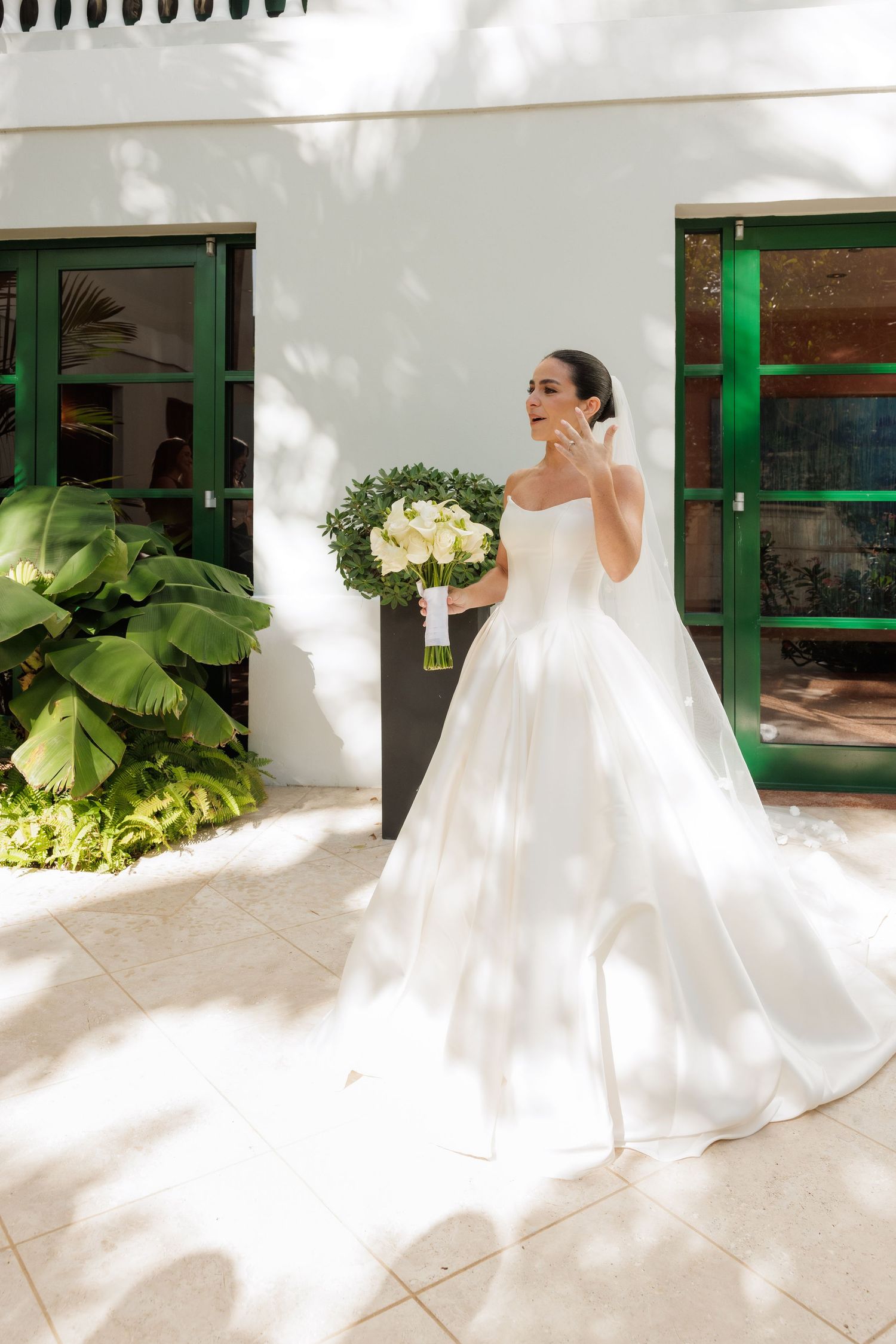 A bride in a white satin gown holds a bouquet of cream flowers against a white building with green accents.
