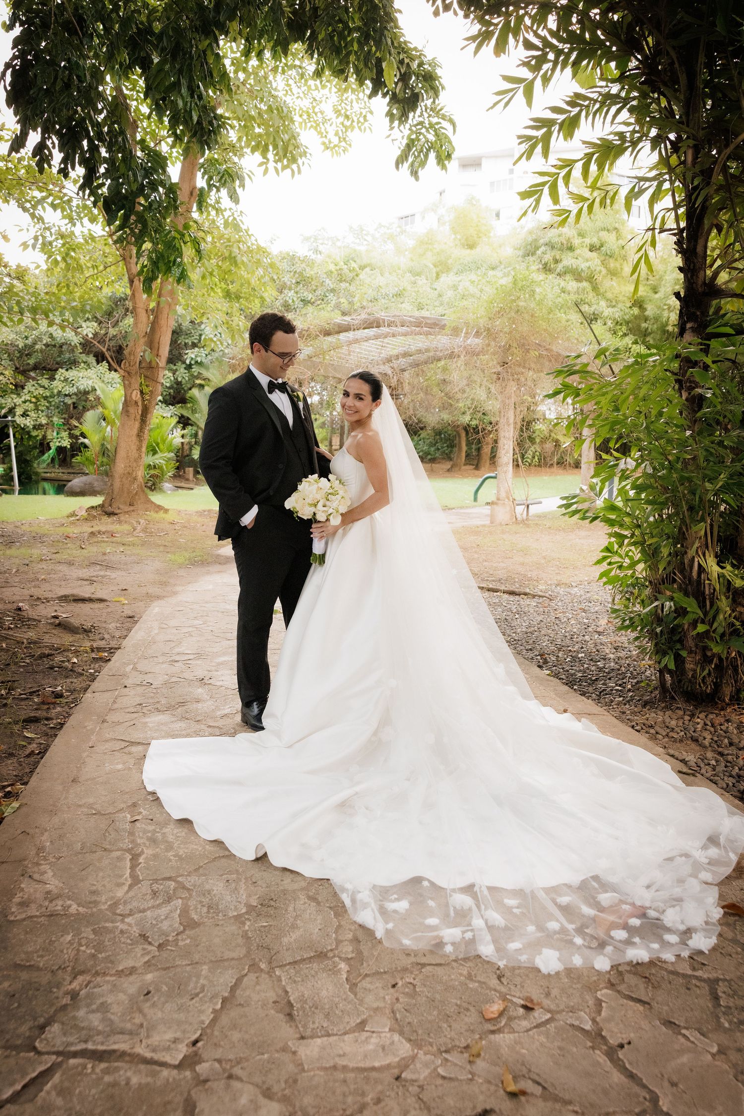 A wedding couple poses on a stone path surrounded by lush greenery and natural lighting.