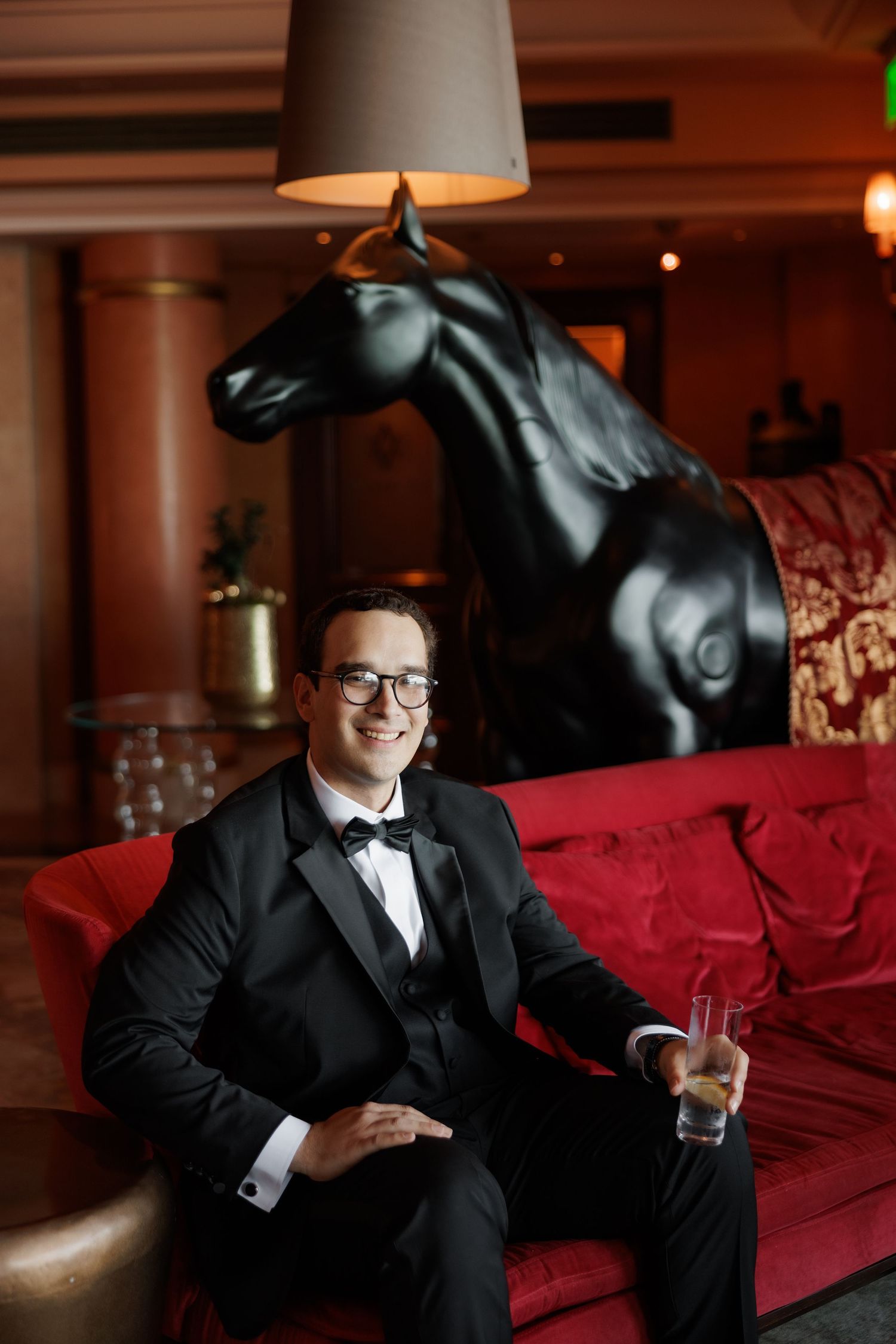 Person in formal black suit sitting on red couch with cocktail glass in moody lighting.