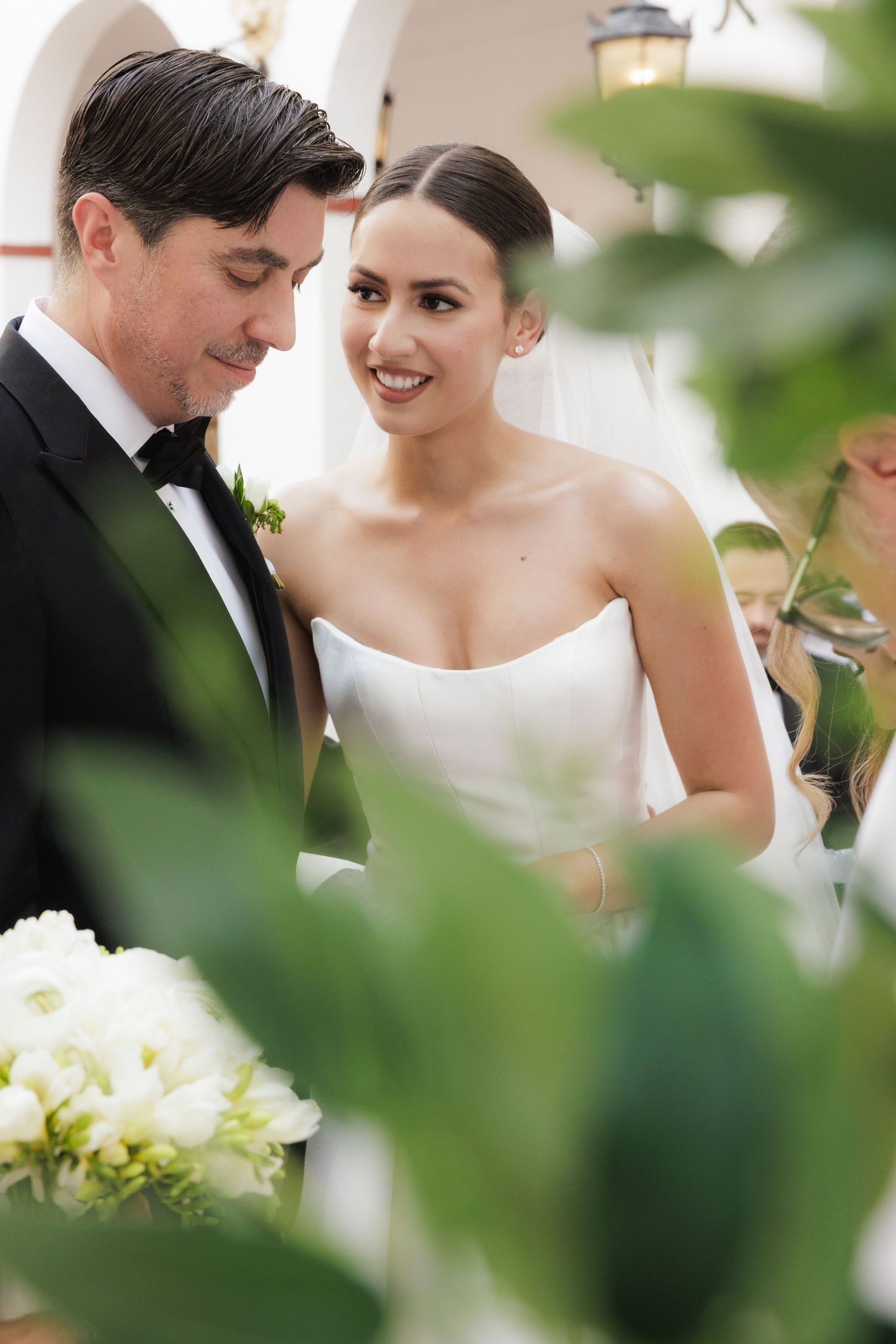 Elegant couple in formal wedding attire share intimate moment among greenery and white flowers.