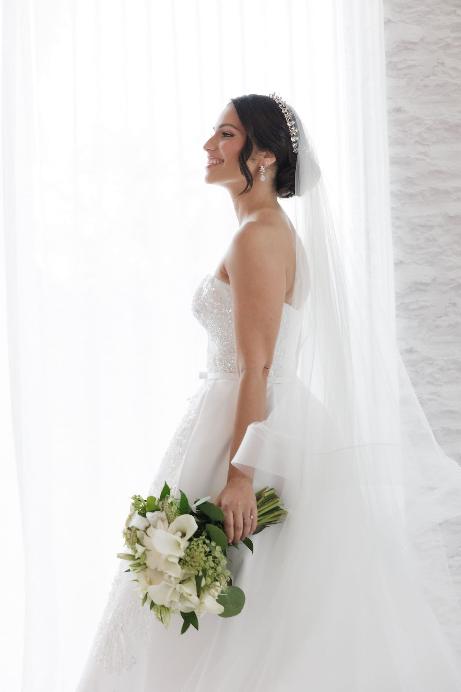A bride in a strapless wedding gown holds a white floral bouquet against sheer white curtains.