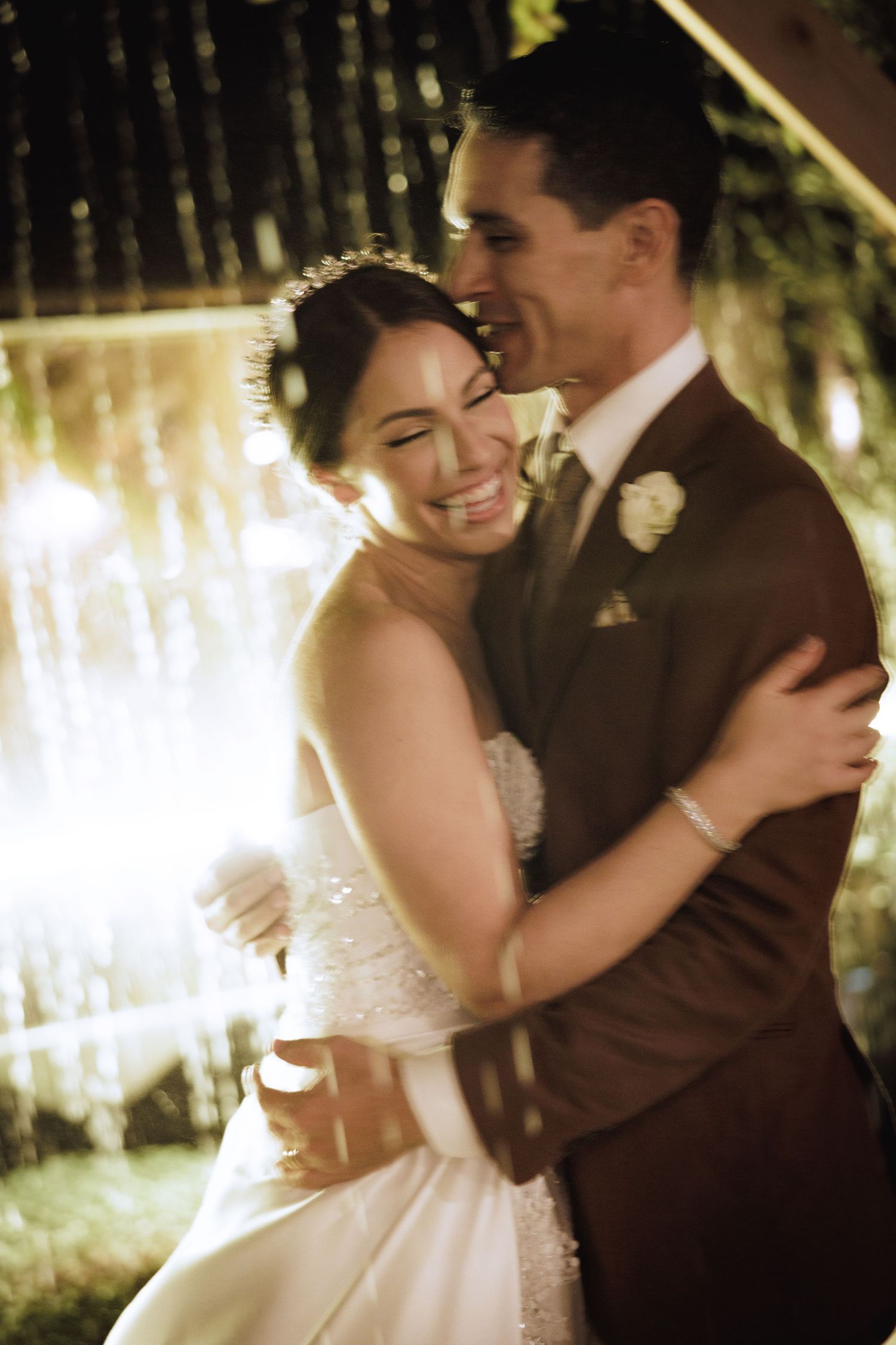 A joyful wedding couple embraces and laughs together in a romantic outdoor setting with twinkling lights.