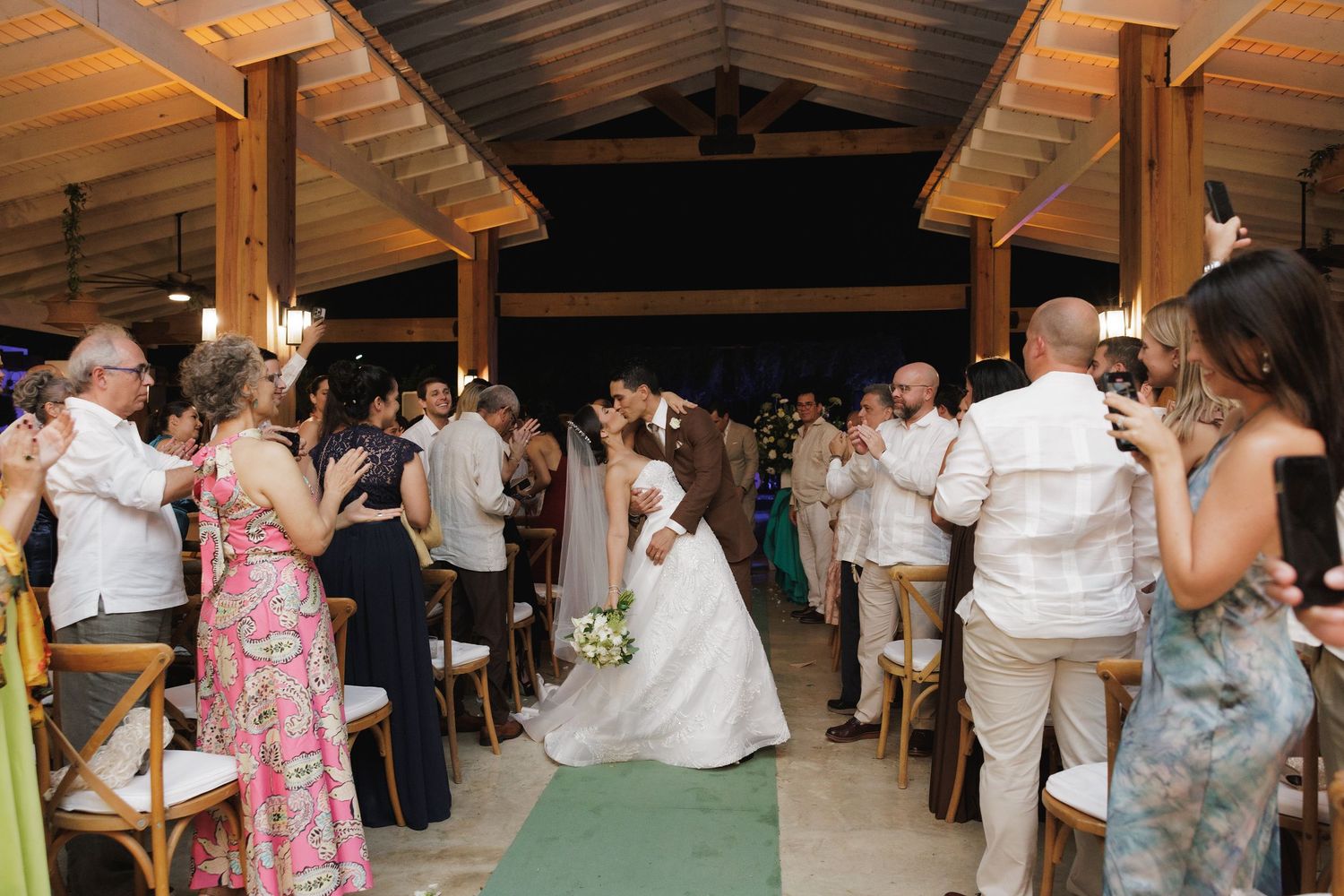 Wedding guests celebrate during a reception under a wooden pavilion with string lights.
