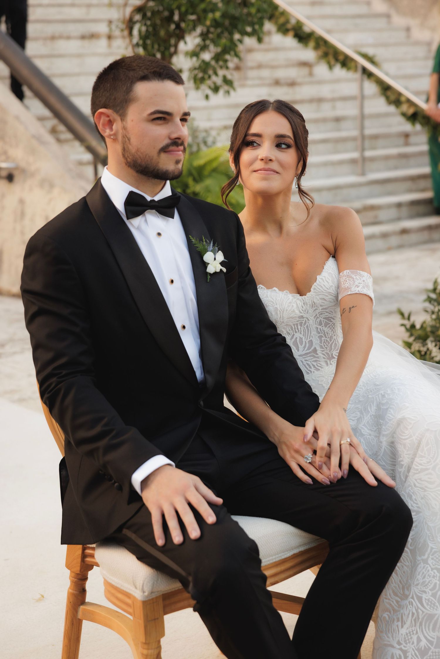 Elegant couple seated together on wooden chairs during formal outdoor event.
