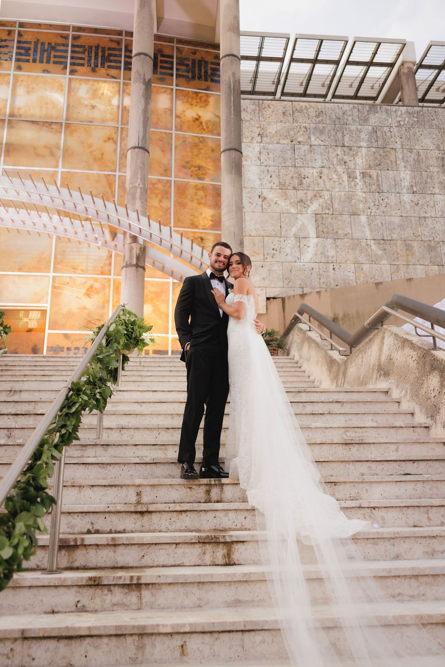 Newlyweds share an intimate moment on elegant stone steps with sunlit architecture in background.
