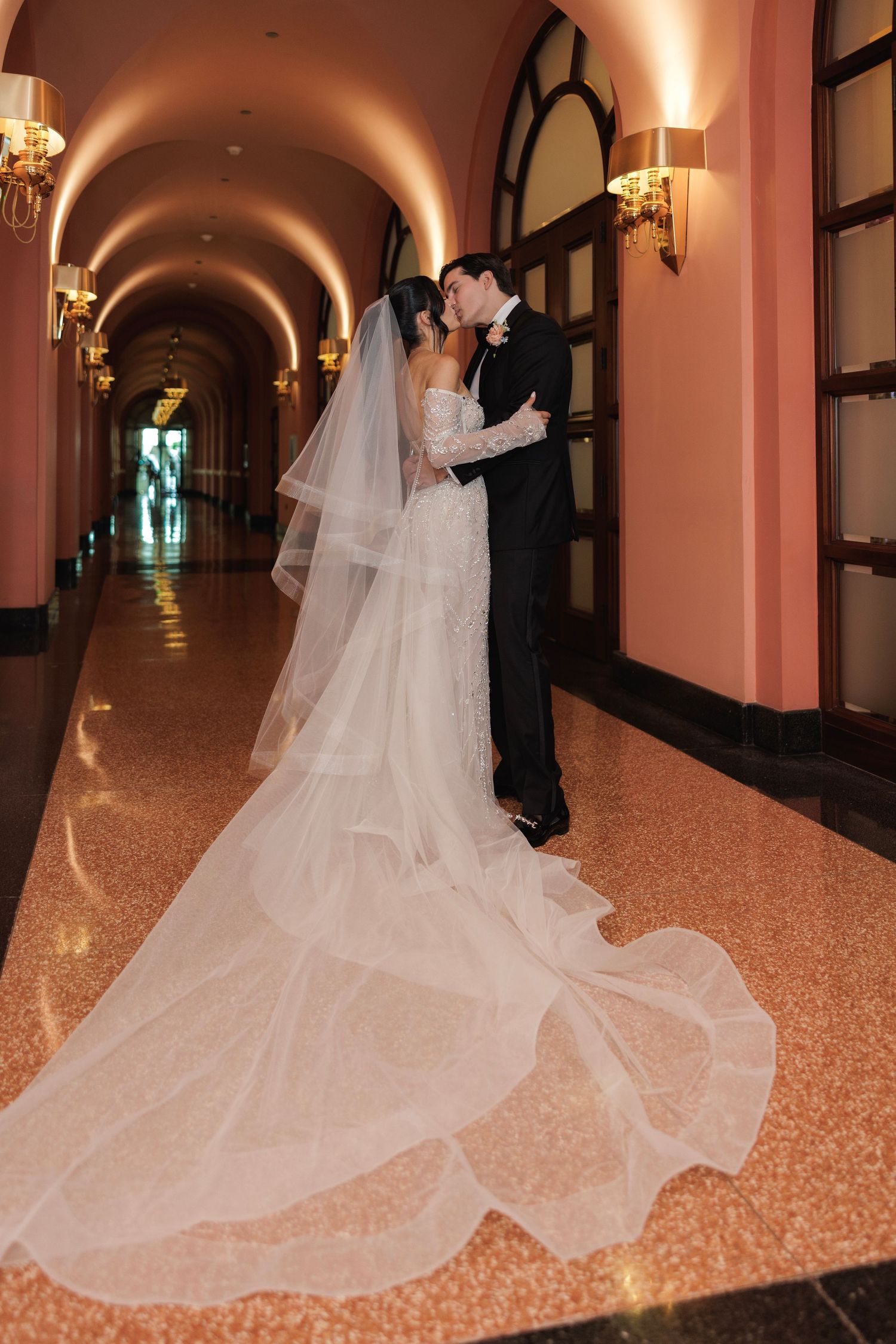 A wedding couple shares an intimate moment in a grand hallway as the bride's long veil trails behind them.