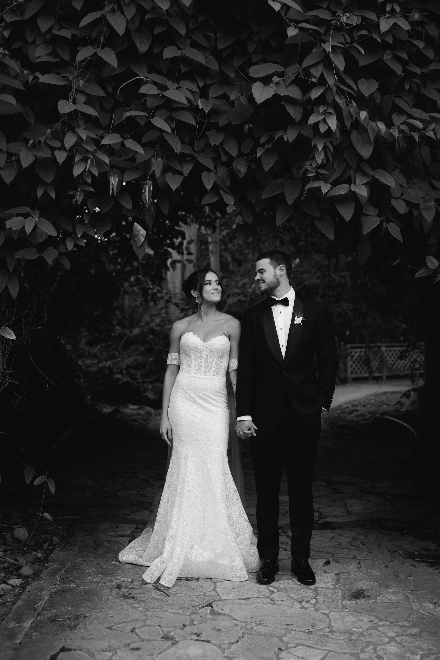 An elegant black and white portrait of newlyweds standing beneath lush foliage on a stone path.