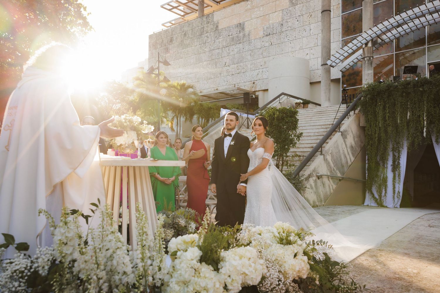 A magical outdoor wedding ceremony illuminated by golden sunlight at a stone venue with white floral decorations.