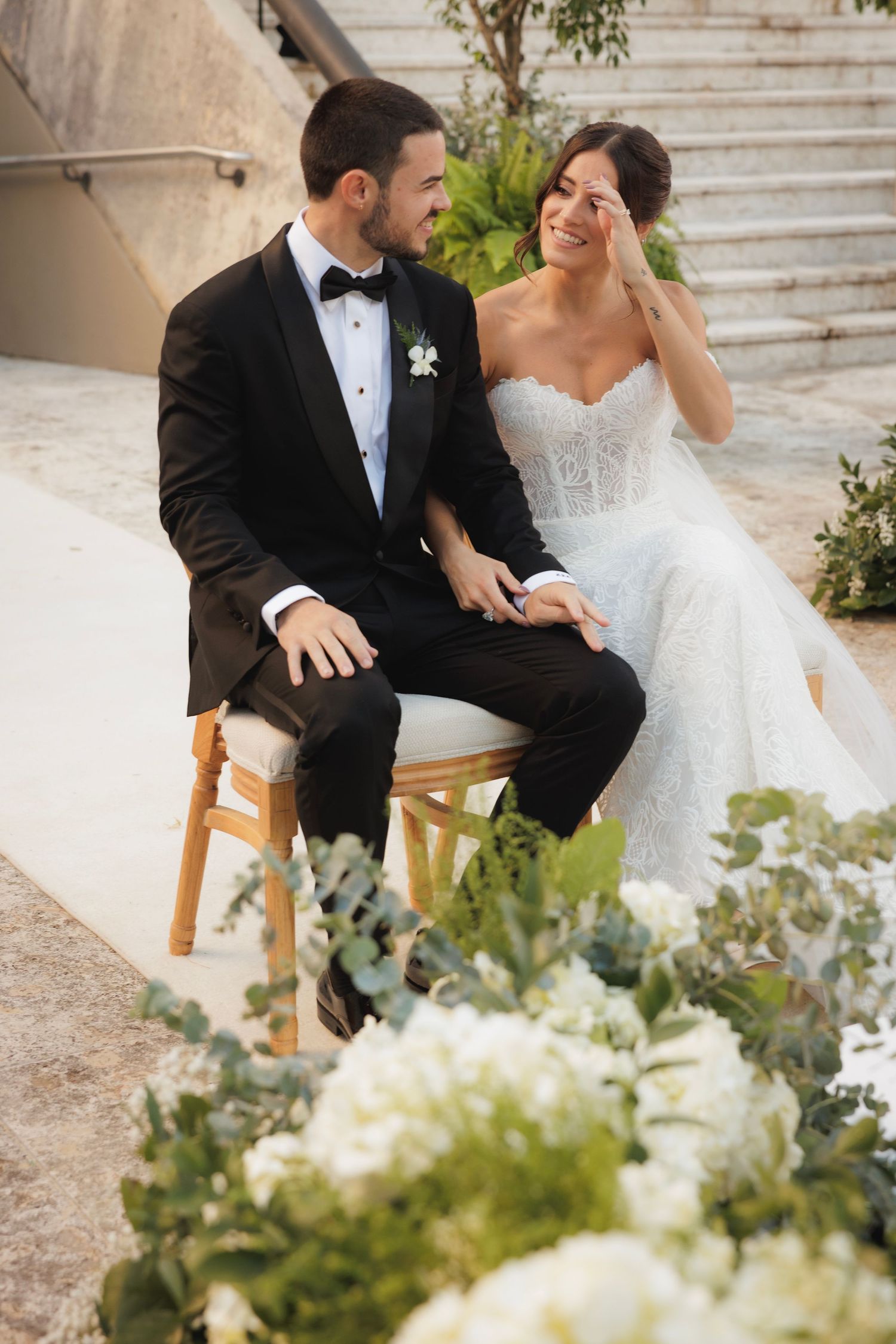 A couple shares an intimate moment seated together amid white floral decorations at an outdoor wedding venue.