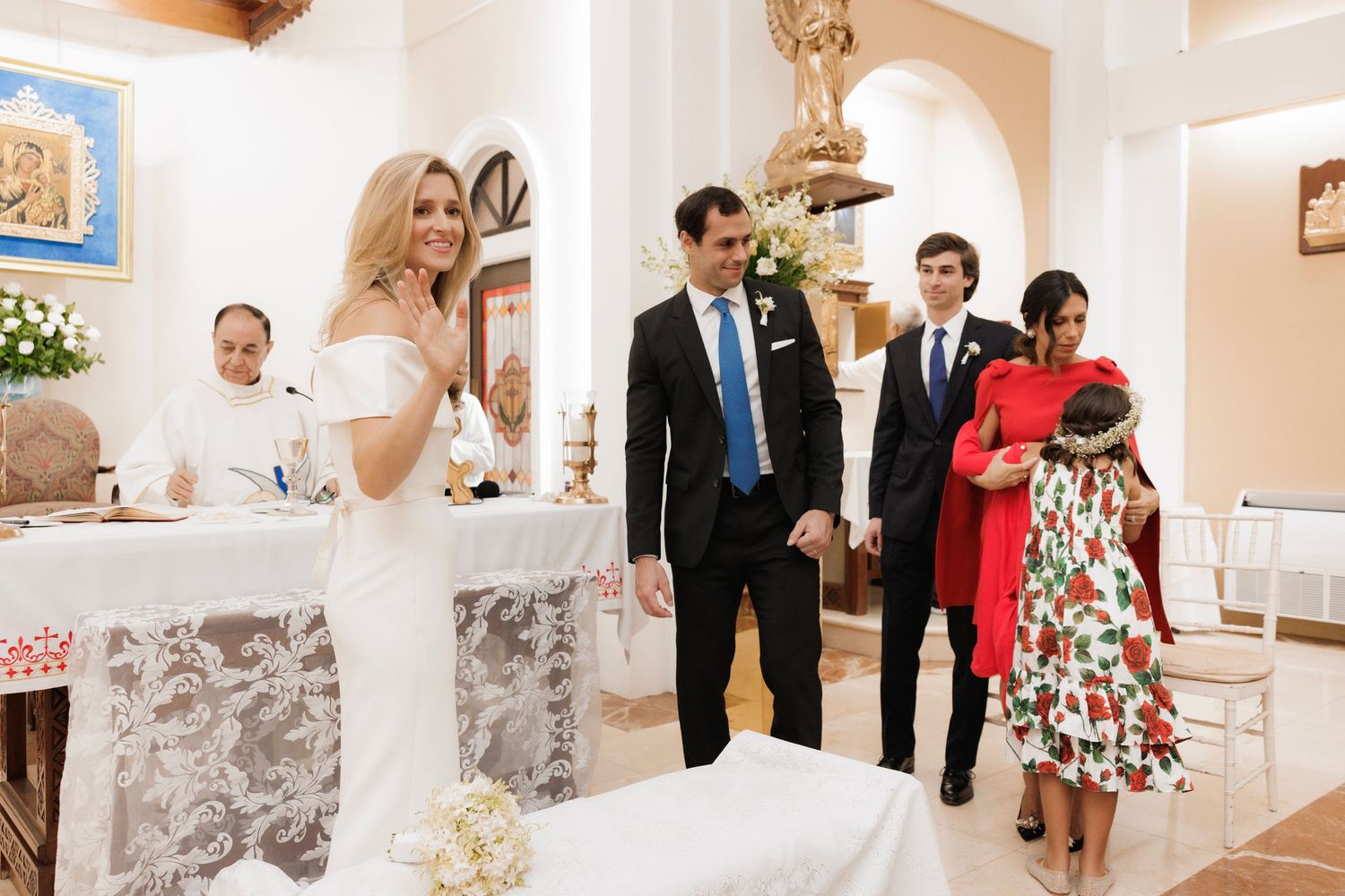 Wedding guests stand in an ornate white chapel interior decorated with floral arrangements and arches.