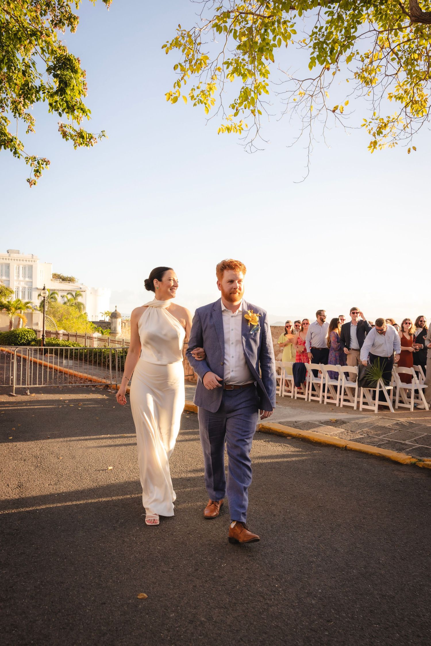 A couple walks along a sunlit path with autumn leaves overhead during their outdoor wedding celebration.