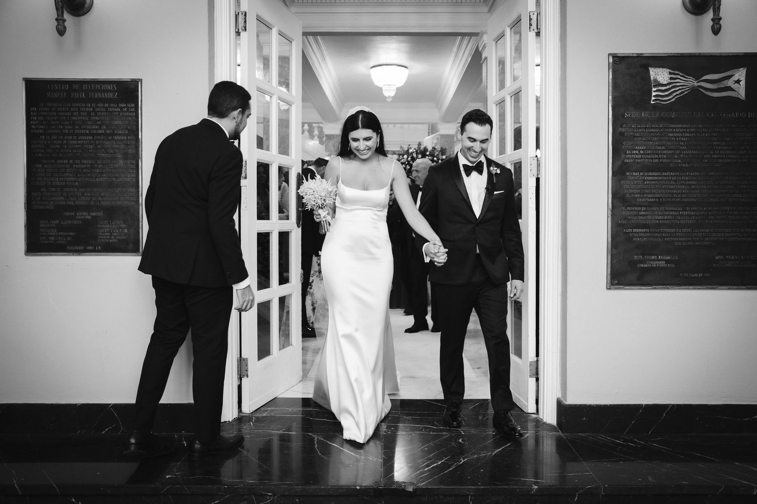 Black and white photo of a wedding couple walking through an elegant hallway with classic architecture.