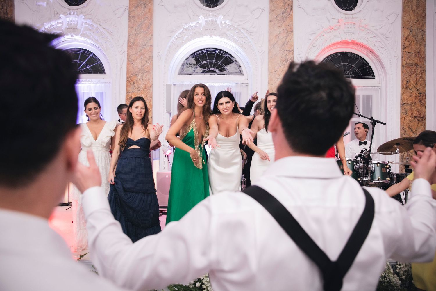 Wedding guests enjoying a lively celebration in a ballroom with arched windows and decorative lighting.