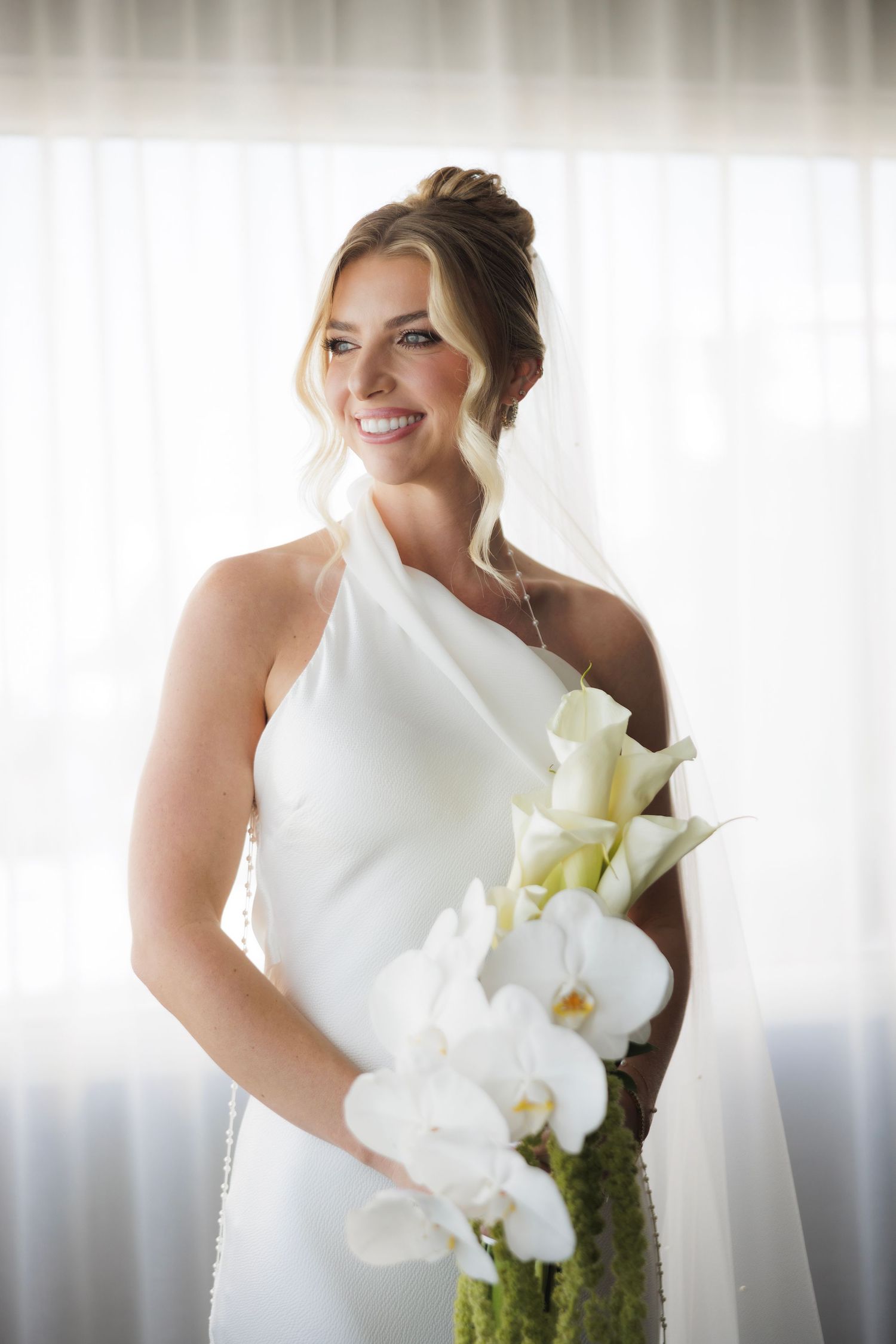A bride in an elegant white one-shoulder dress holds a beautiful bouquet of white orchids and calla lilies.