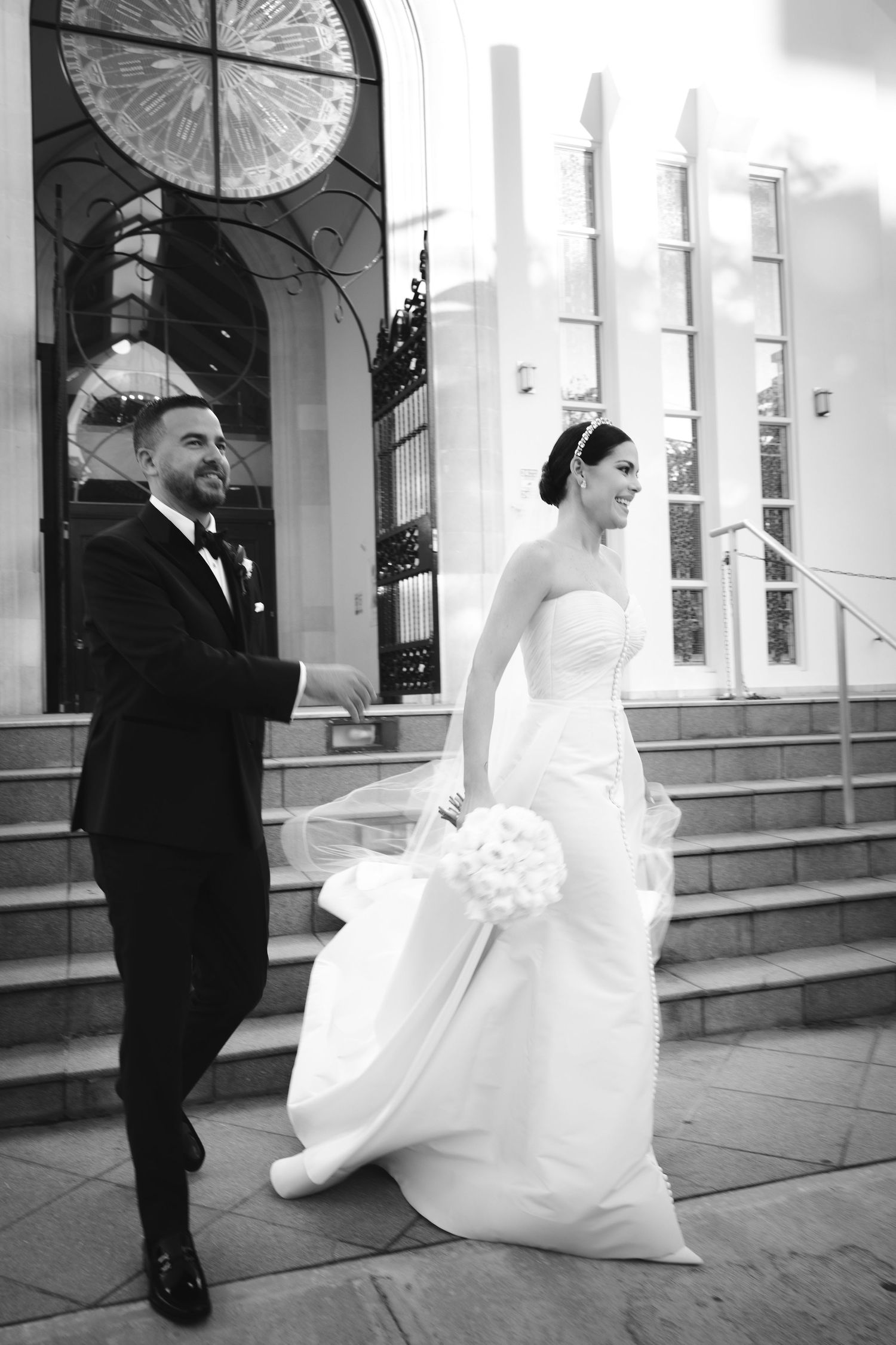 A black and white wedding photograph showing a bride in a white gown and groom in a suit descending church steps.