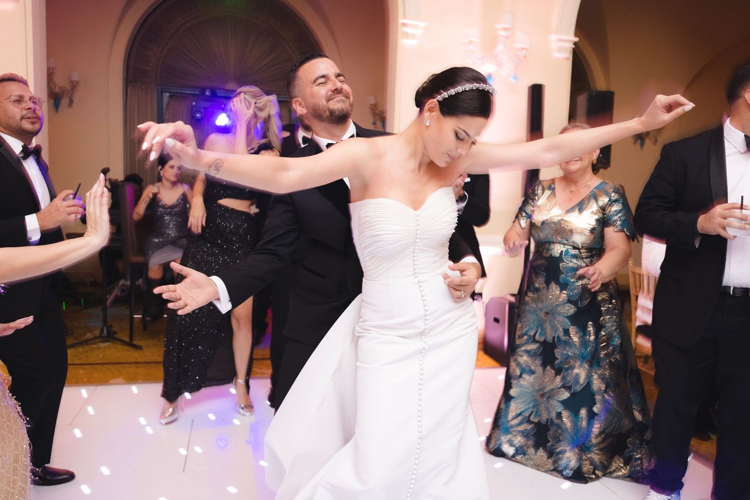 Wedding guests dance together at a reception with people in formal attire spreading their arms in celebration.