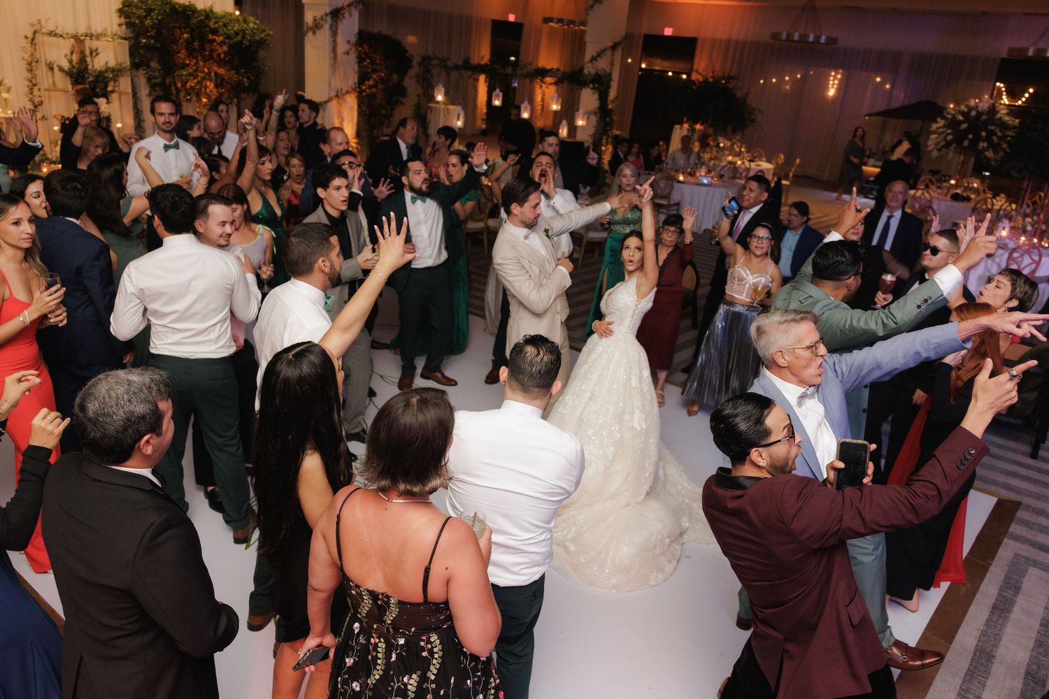 Wedding guests enjoy dancing on a crowded reception dance floor with festive lighting and decorations.
