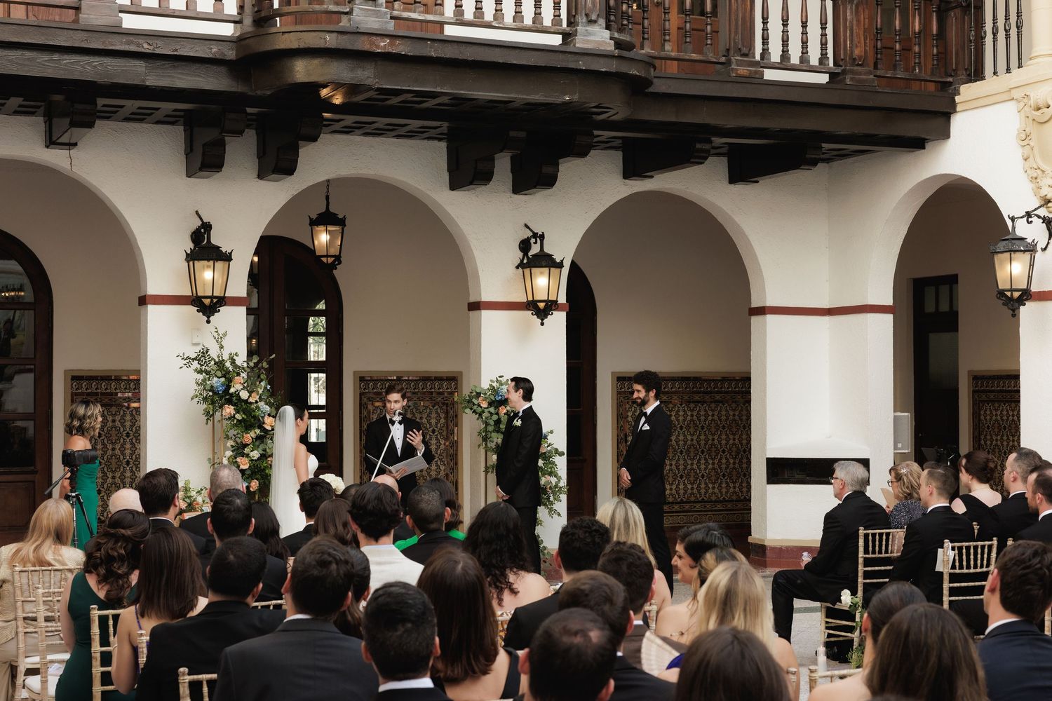 People gather in an elegant open-air courtyard with white arches and columns for an event or gathering.