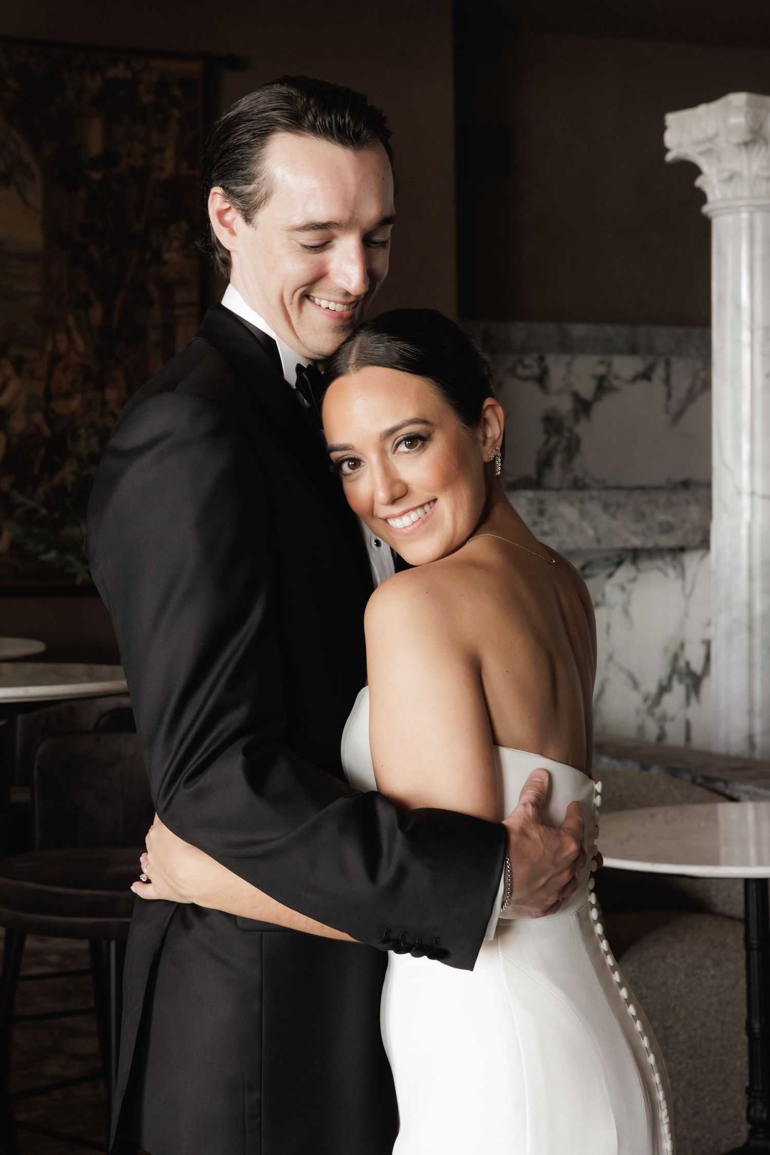 Happy newlyweds embrace in formal wedding attire inside an elegant venue with marble and classical decor.