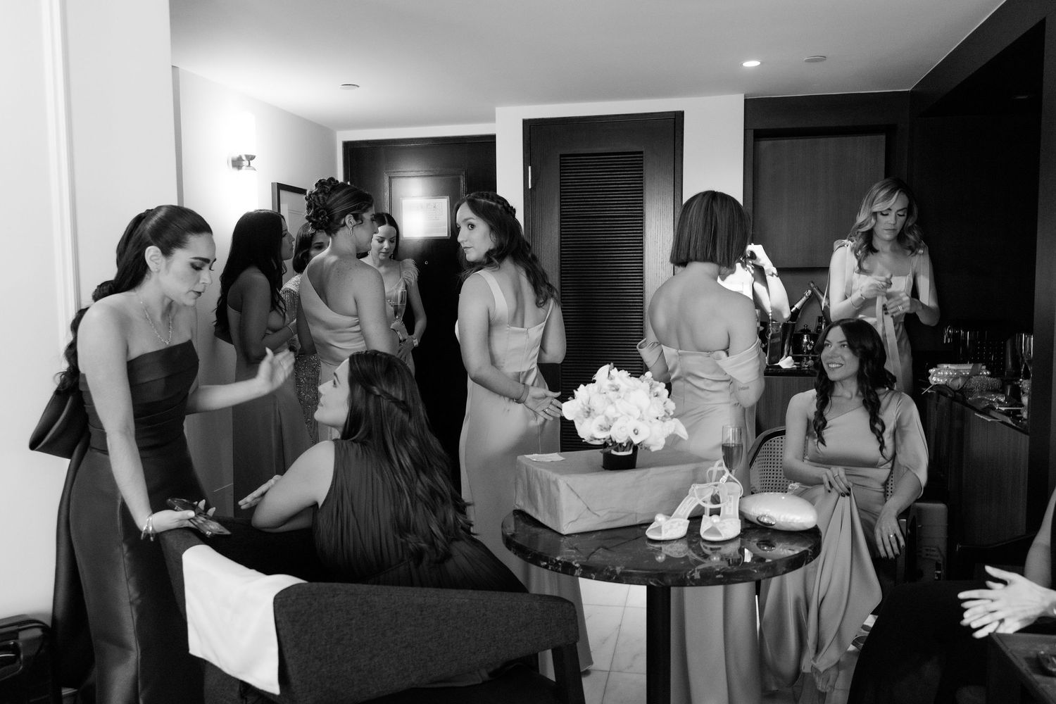 A candid black and white photo showing a group getting ready in a room with a gift box and flowers on the table.